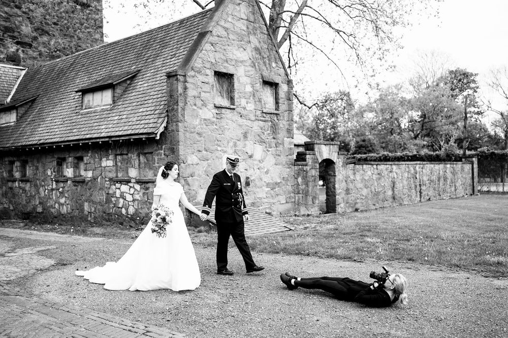 Bride and groom walking past a photographer lying on the ground at a stone building wedding venue.