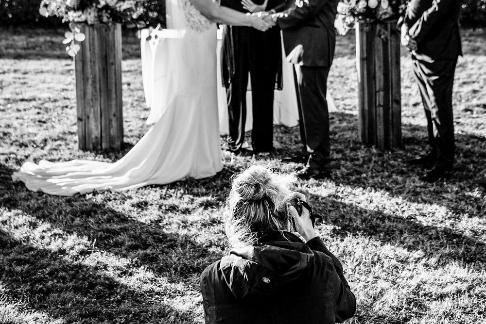 Wedding photographer kneeling on grass to capture bride and groom during outdoor ceremony in black and white.