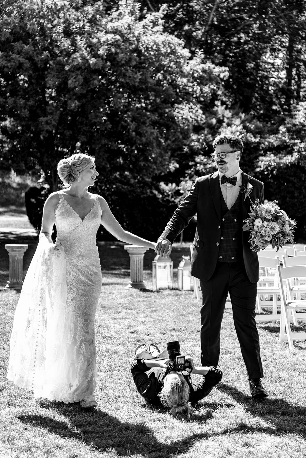 Wedding photographer lying on grass to capture bride and groom holding hands at outdoor ceremony.