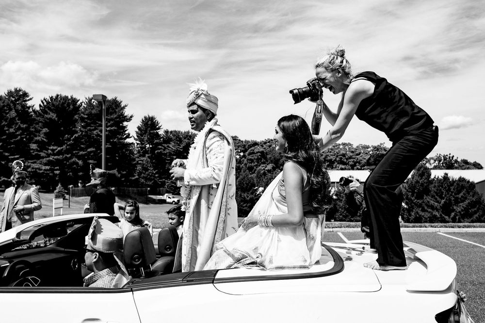 Photographer standing on car hood to photograph bride and groom in convertible at outdoor wedding.