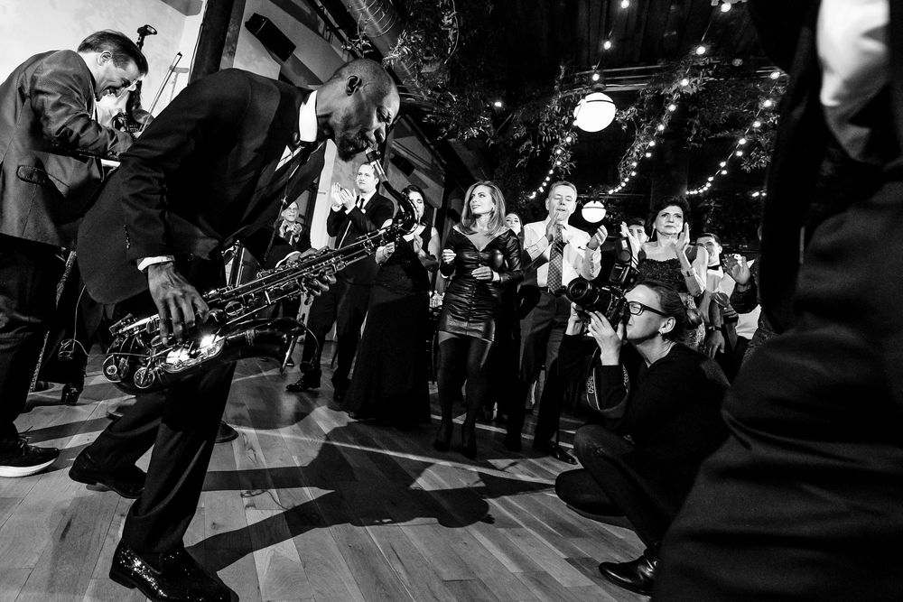 Wedding photographer crouching to capture guests dancing near saxophonist at lively reception in black and white.