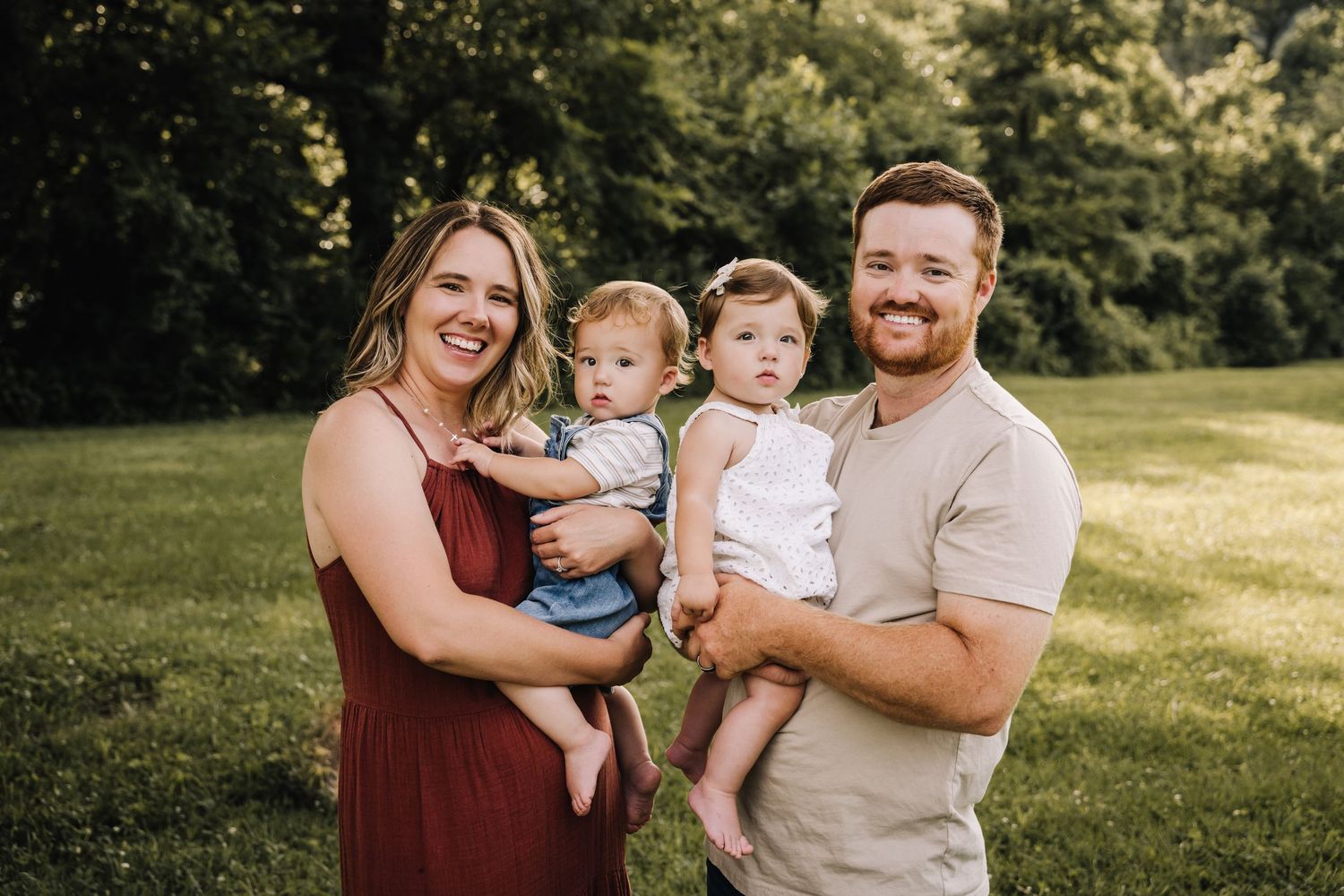 A family stands together outdoors on a sunny day in a grassy area with trees in the background. Family photographer in Middle Tennessee capt