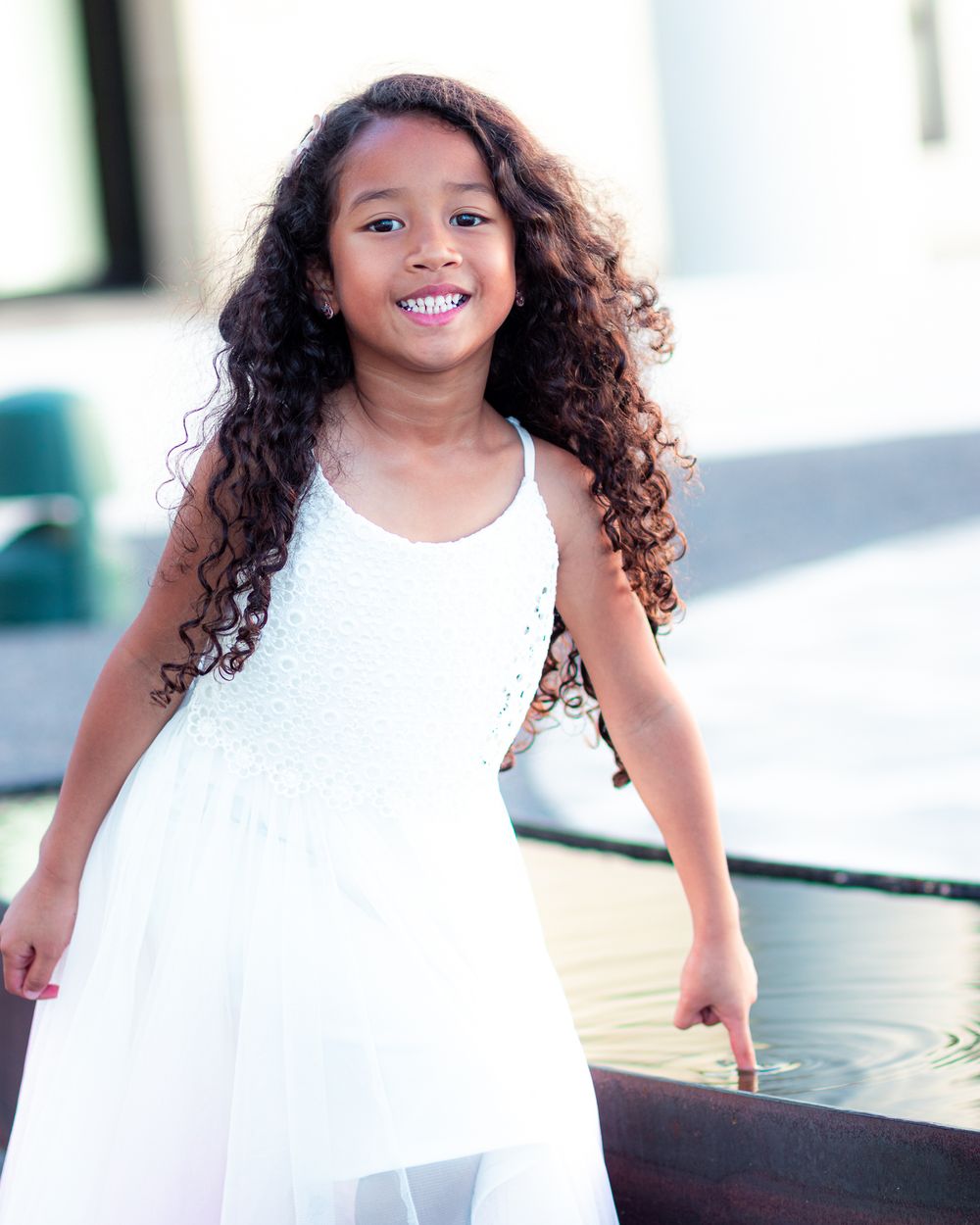 Young child in white dress twirls and smiles while playing in bright indoor setting.