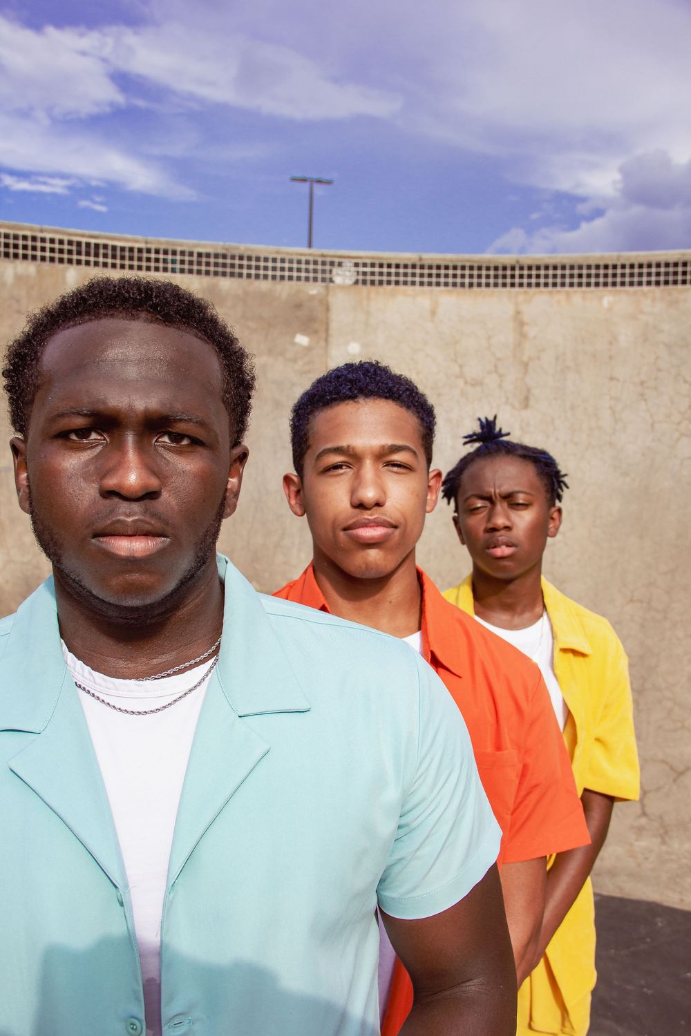 Group of people wearing colorful clothing standing against a concrete wall under blue sky.
