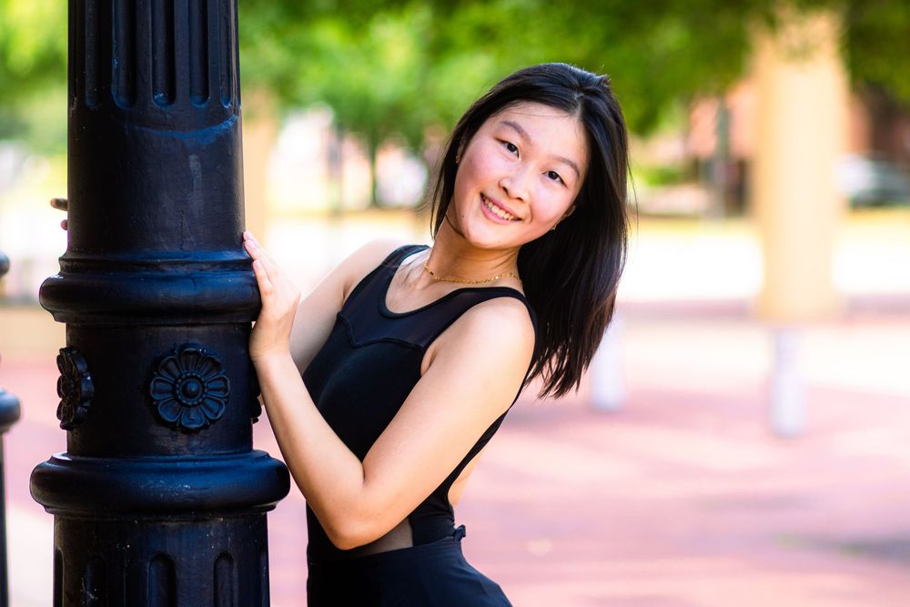 Person in a black top poses playfully against a black lamppost in an outdoor setting with blurred background.