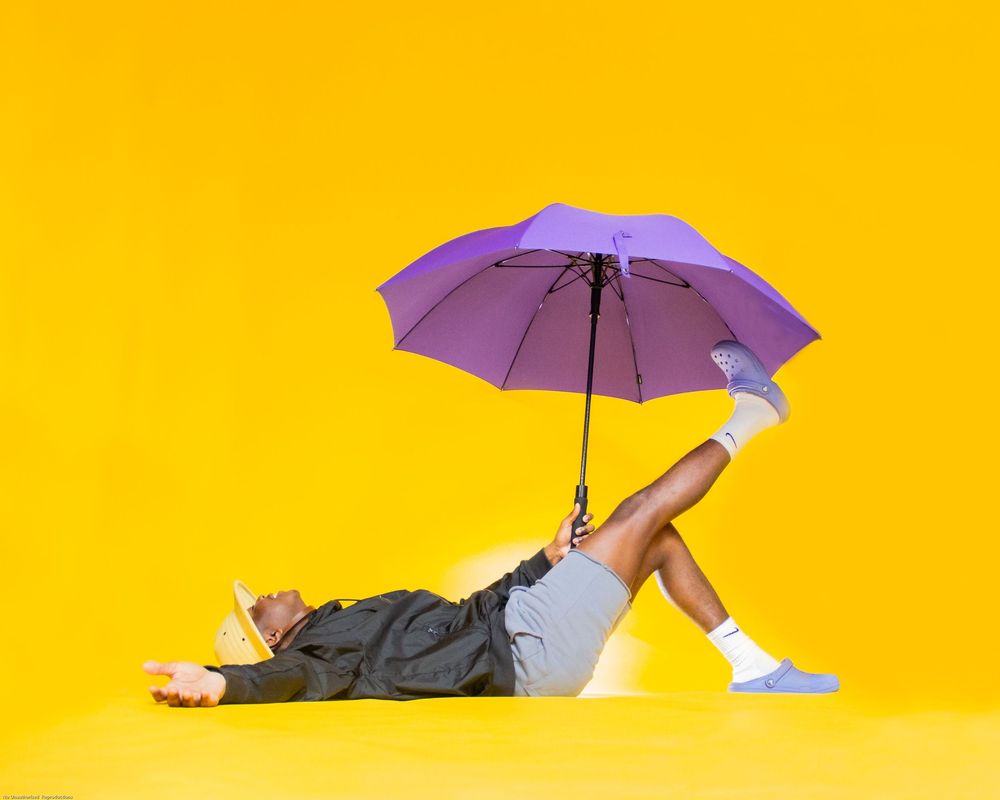A person floating horizontally with a purple umbrella against a bright yellow background in a surreal pose.