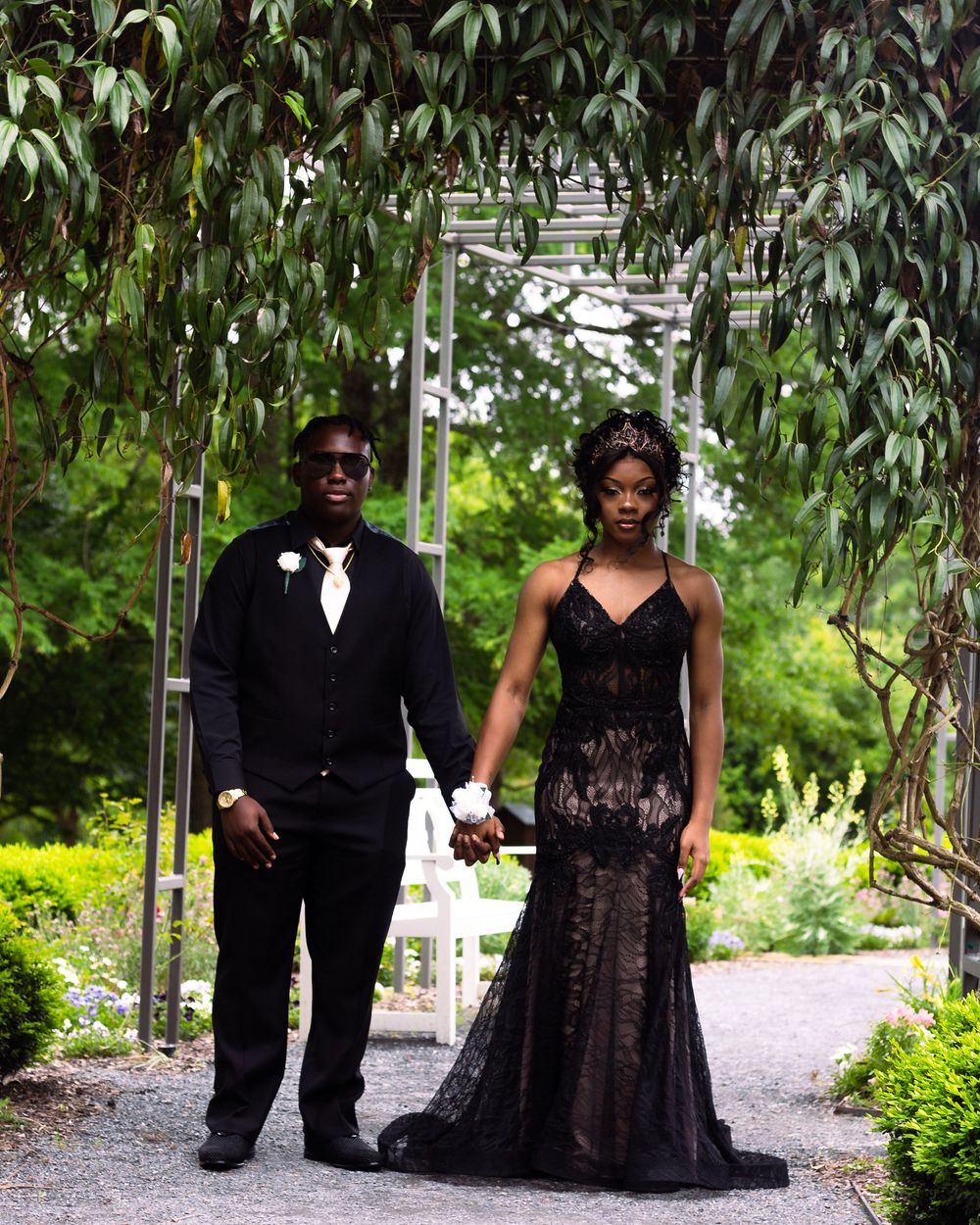 A couple poses together on a garden path surrounded by lush greenery and hanging willow branches.