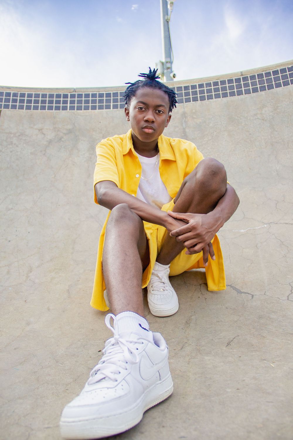 A person in a bright yellow shirt and white sneakers sits casually against a curved concrete wall outdoors.