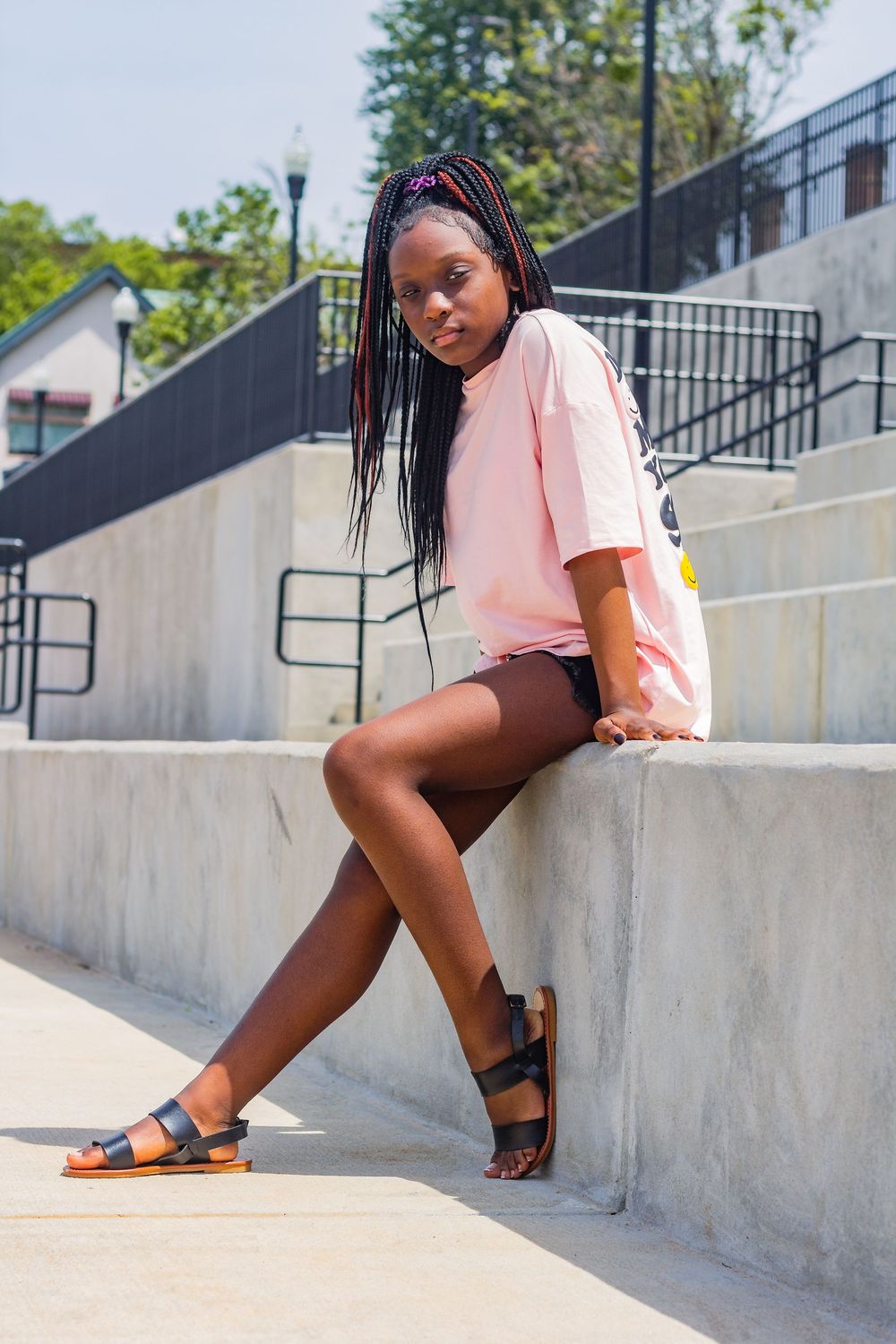 Someone in a casual pink t-shirt and dark sandals sits on concrete steps on a sunny day outdoors.