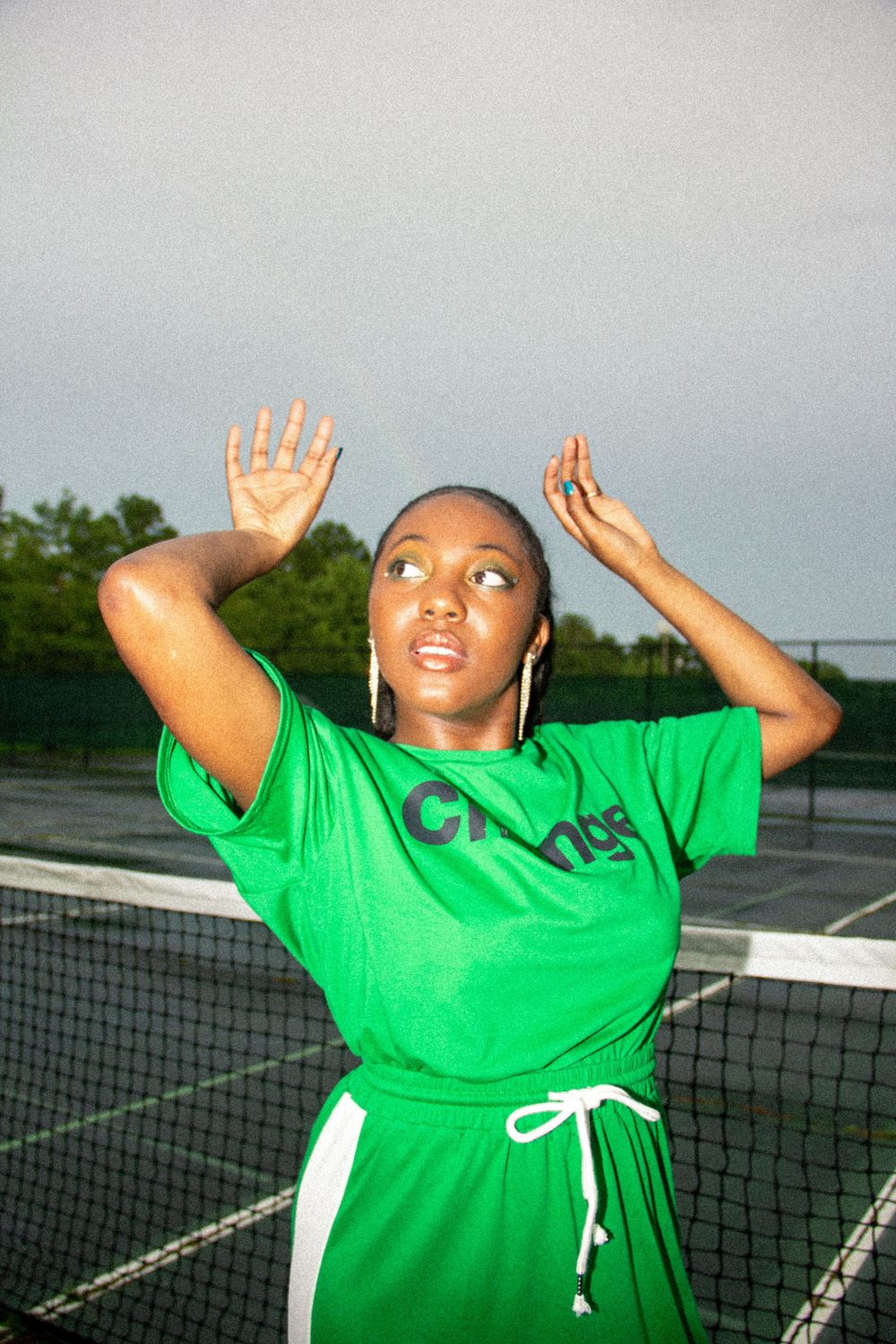 A person in a bright green t-shirt performs a series of expressive dance movements on an outdoor tennis court.