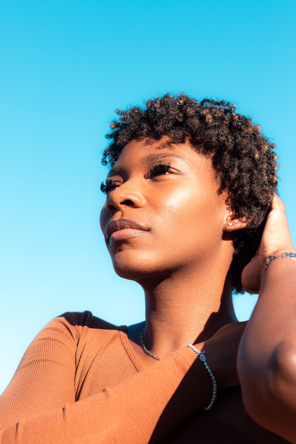 Portrait against vibrant blue sky showing natural curly hair and orange top in warm sunlight.