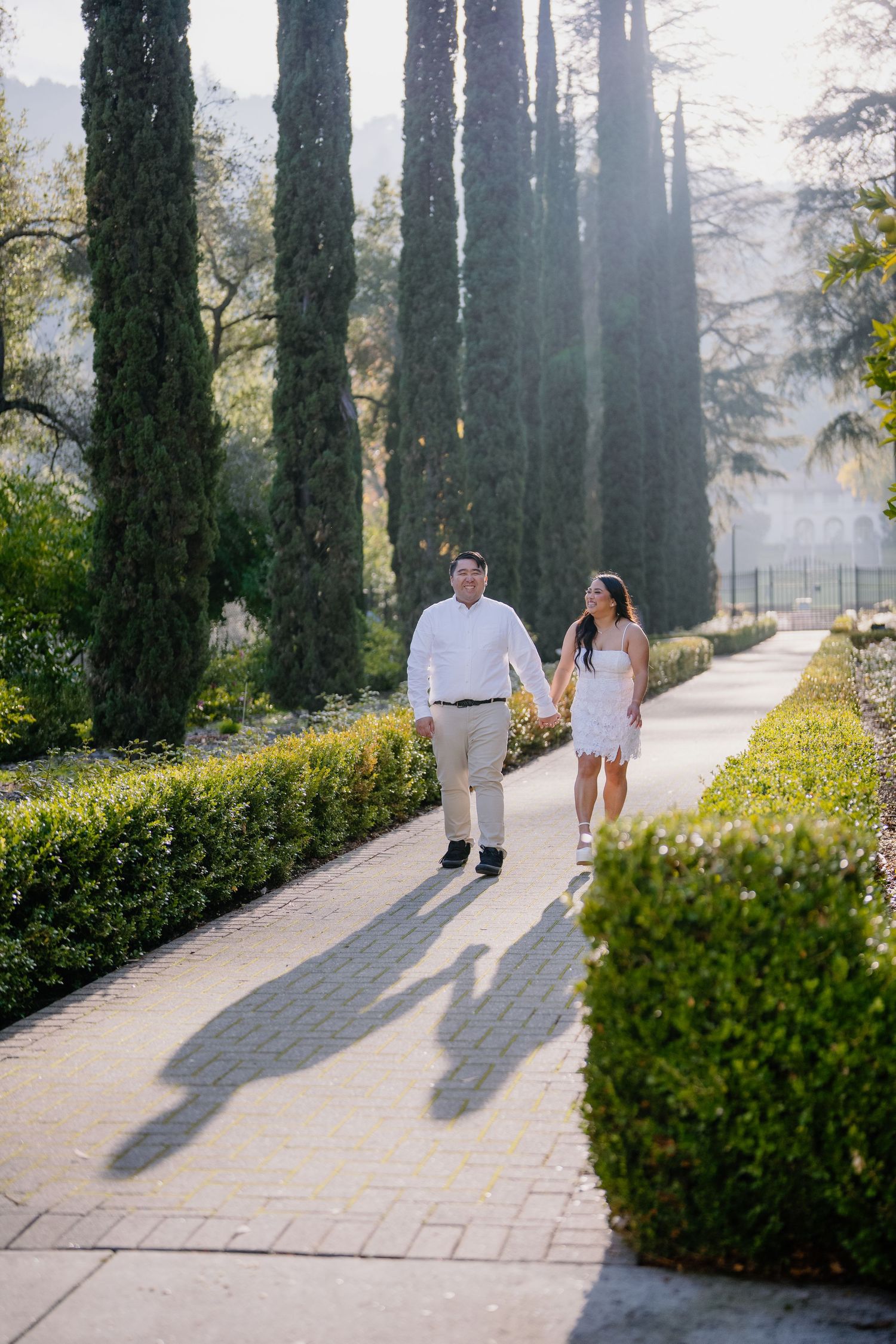A couple walks together on a sunlit garden path lined with tall cypress trees and manicured hedges.