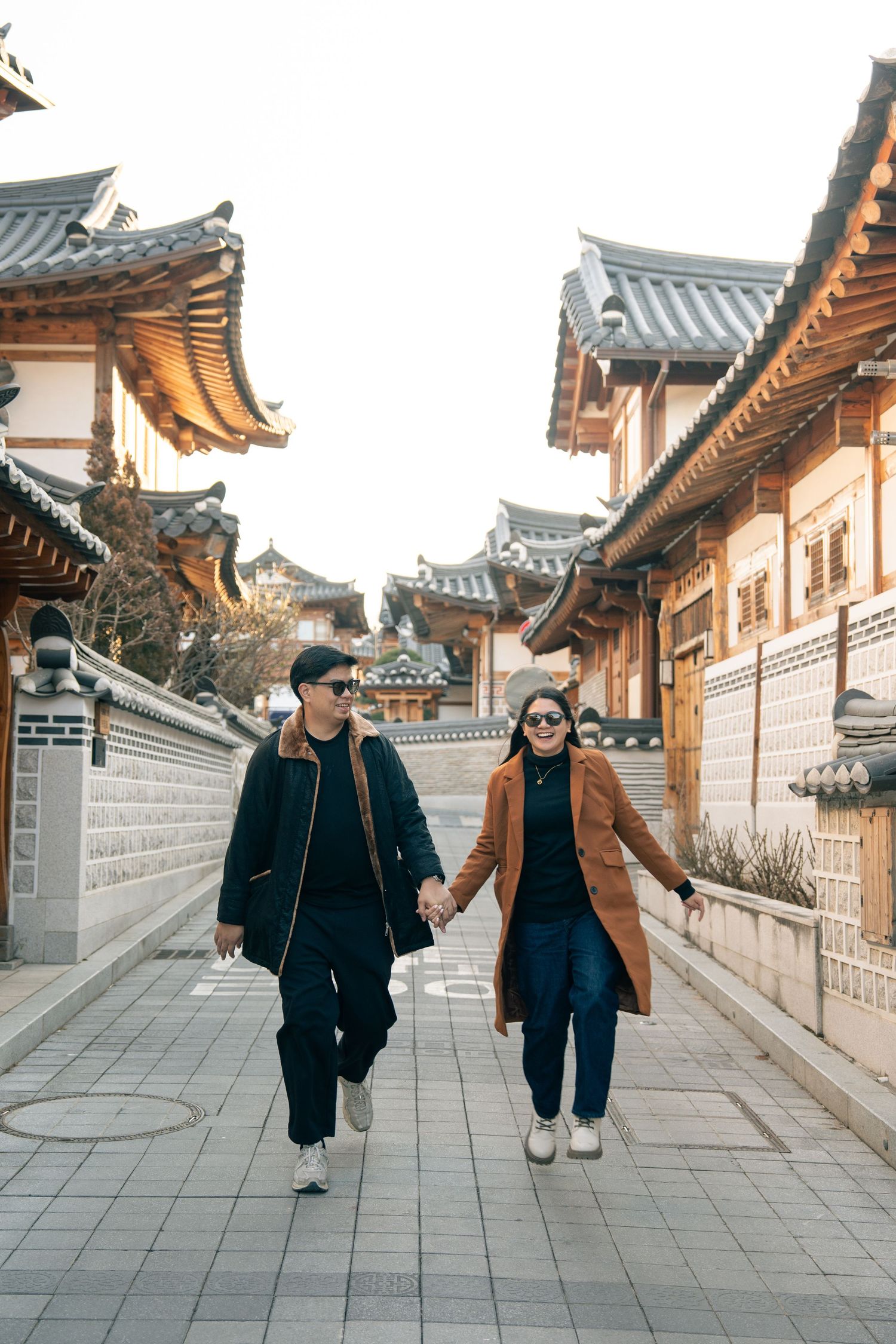 People walking along a traditional Korean hanok village street with stone walls and curved tile roofs in warm sunlight.