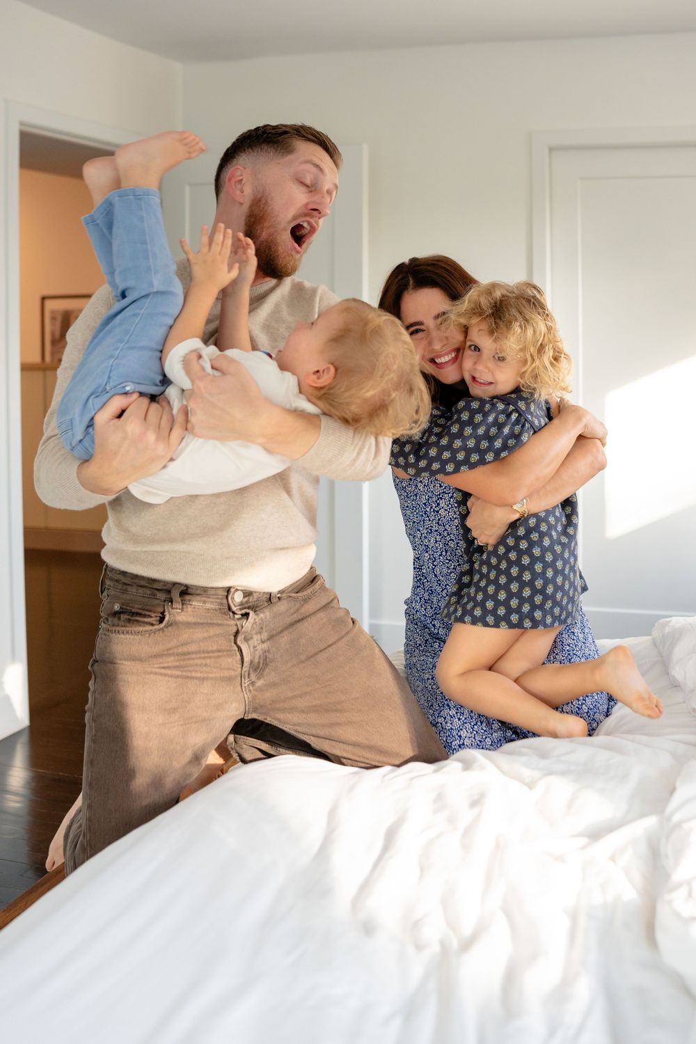 Family having fun playing and laughing together on a white bed in a bright sunny room.