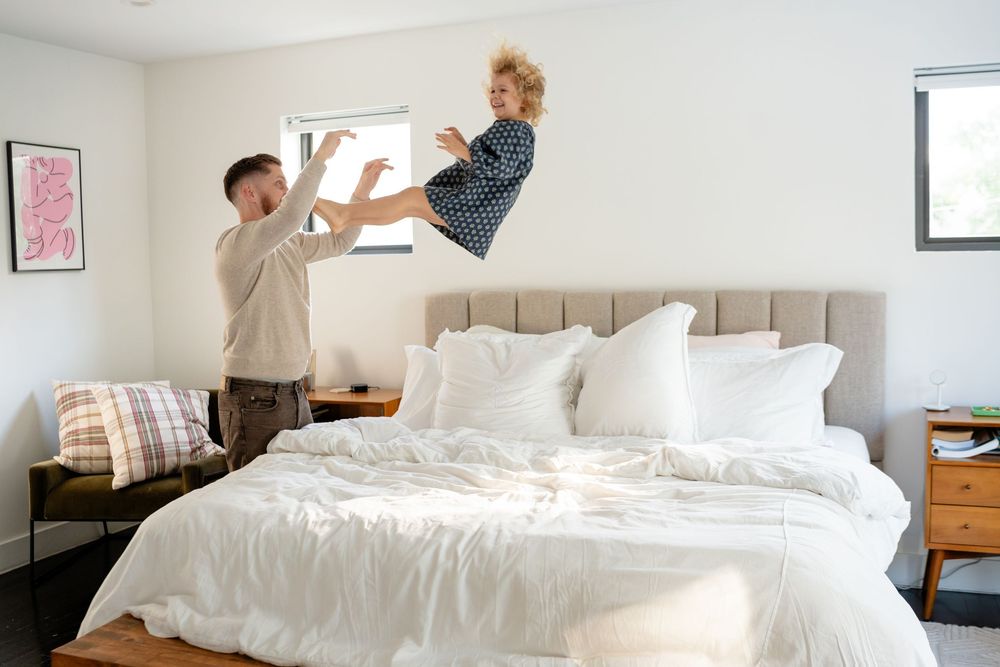 A person making a white bed with fresh linens and pillows in a bright bedroom.