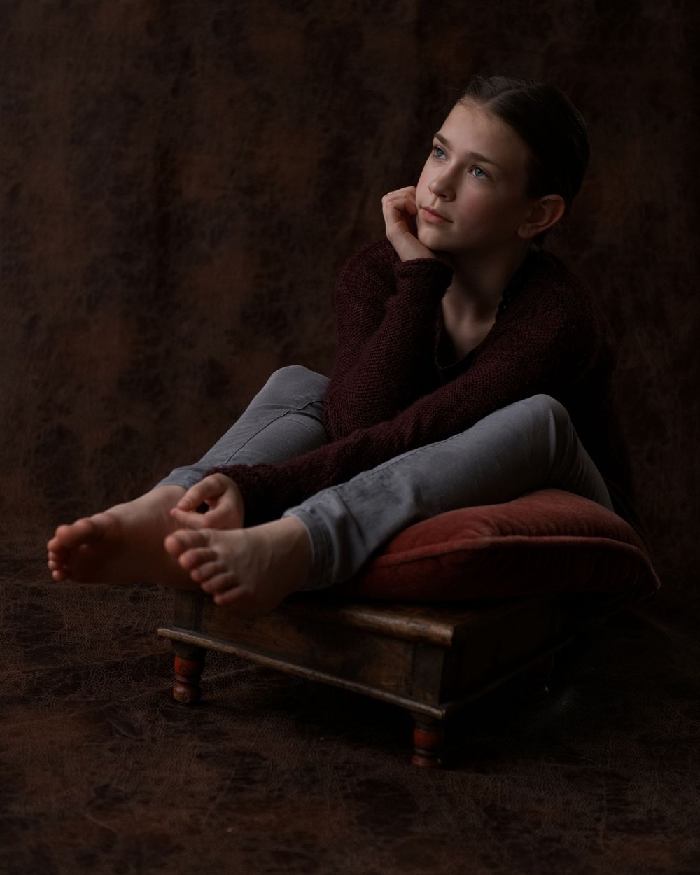 A young girl in a burgundy sweater sits thoughtfully on a wooden stool with a red cushion in a dark studio.