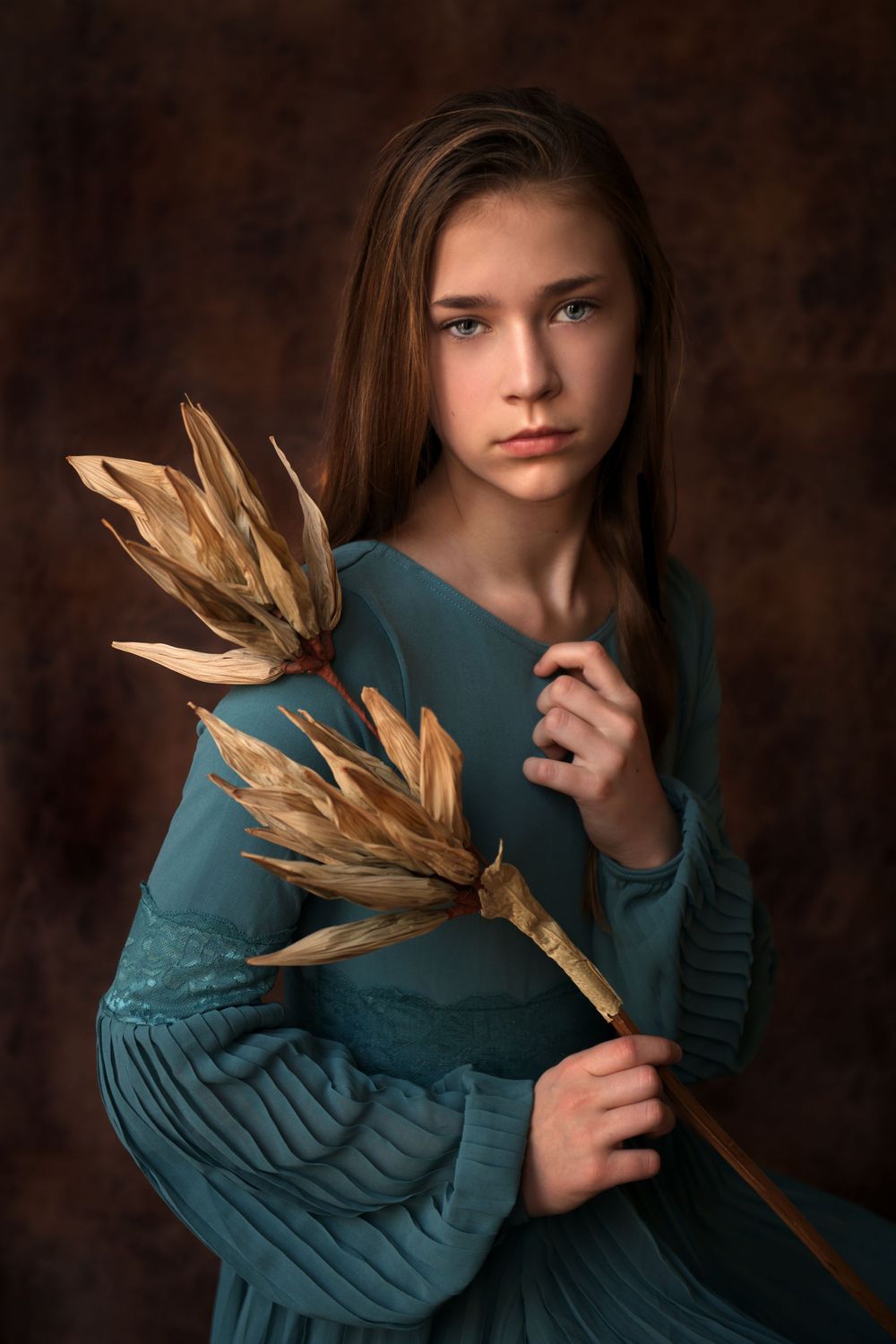 A young girl in a teal dress holds golden dried wheat stalks against a dark brown studio background.