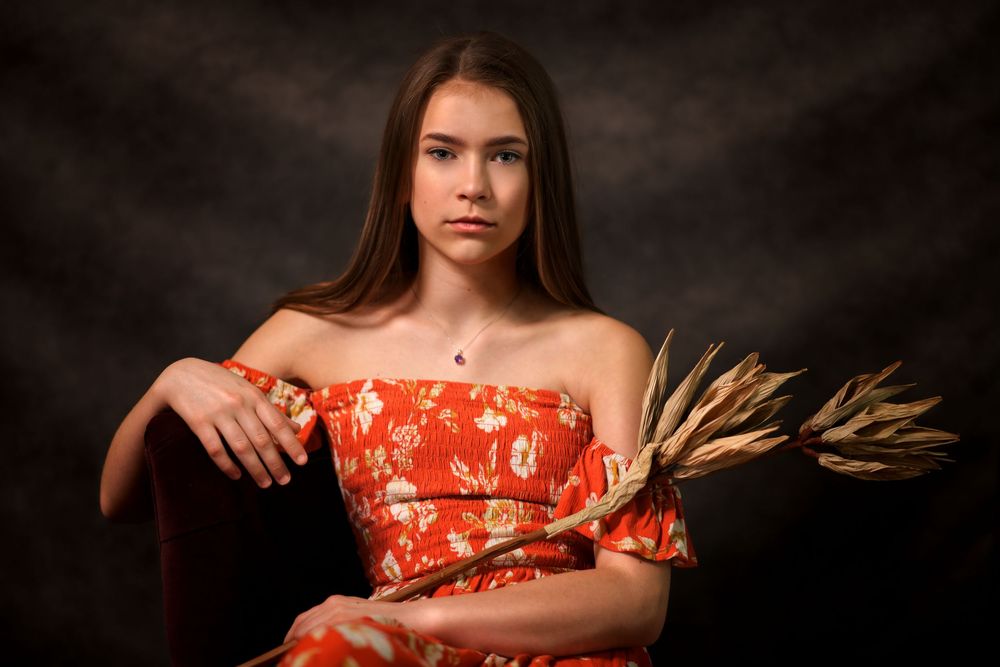 A teenage girl in a red floral off-shoulder dress holds dried wheat while seated in a dark studio setting.