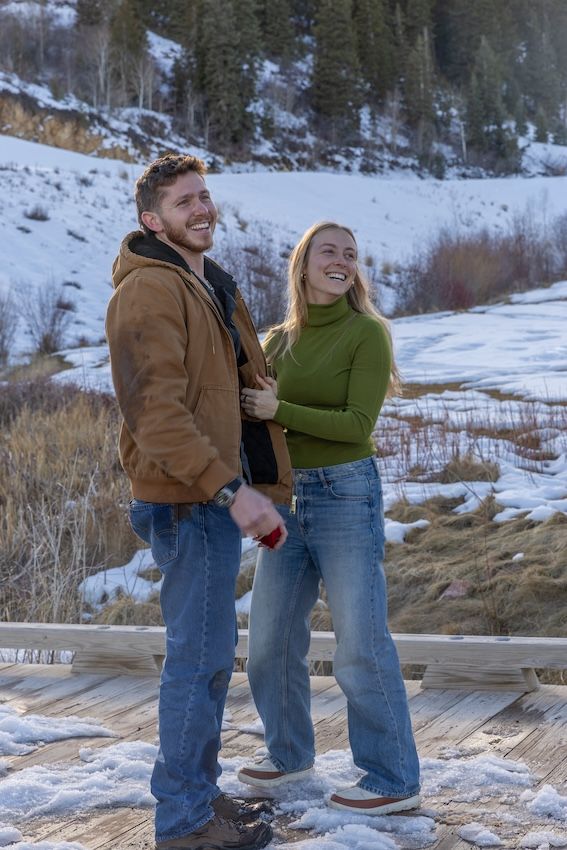 A couple shares a joyful moment on a wooden boardwalk surrounded by snowy winter landscape.