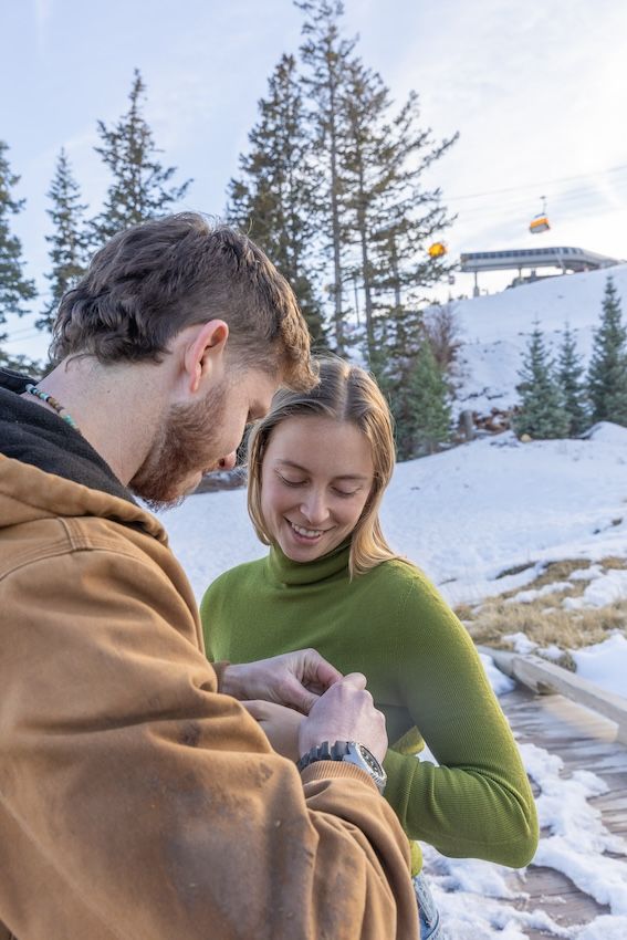 A couple shares a tender moment in winter against a snowy mountain backdrop with evergreen trees.