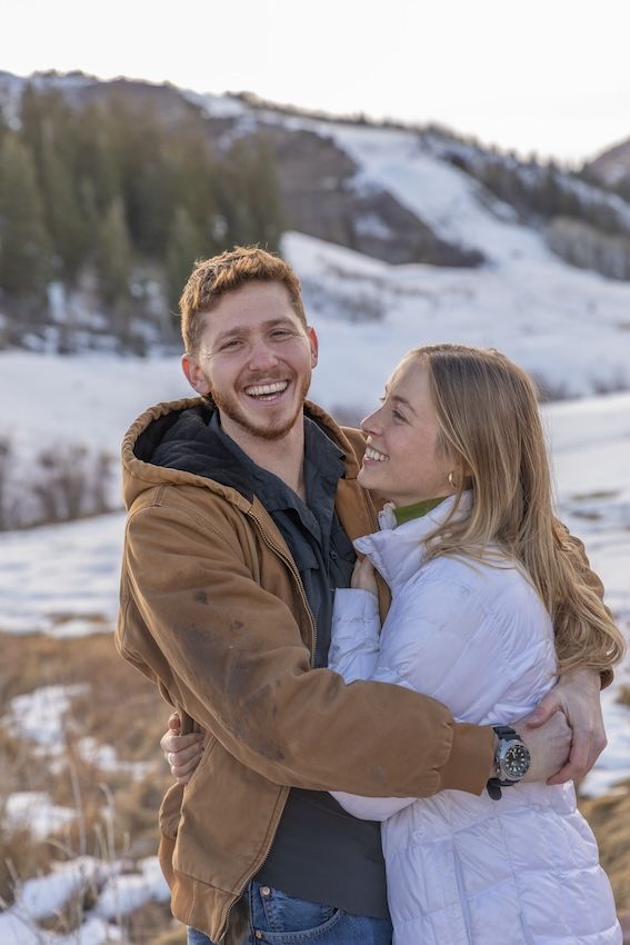 A couple shares joyful embraces and laughter against a scenic snow-covered mountain backdrop.