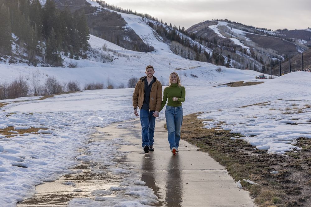 Two people walking along a snowy path with mountains and winter scenery in the background.