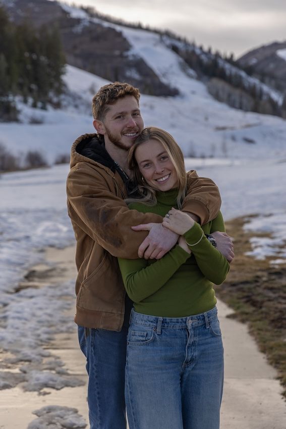 A couple shares a joyful embrace in front of snow-covered mountains on a winter afternoon.