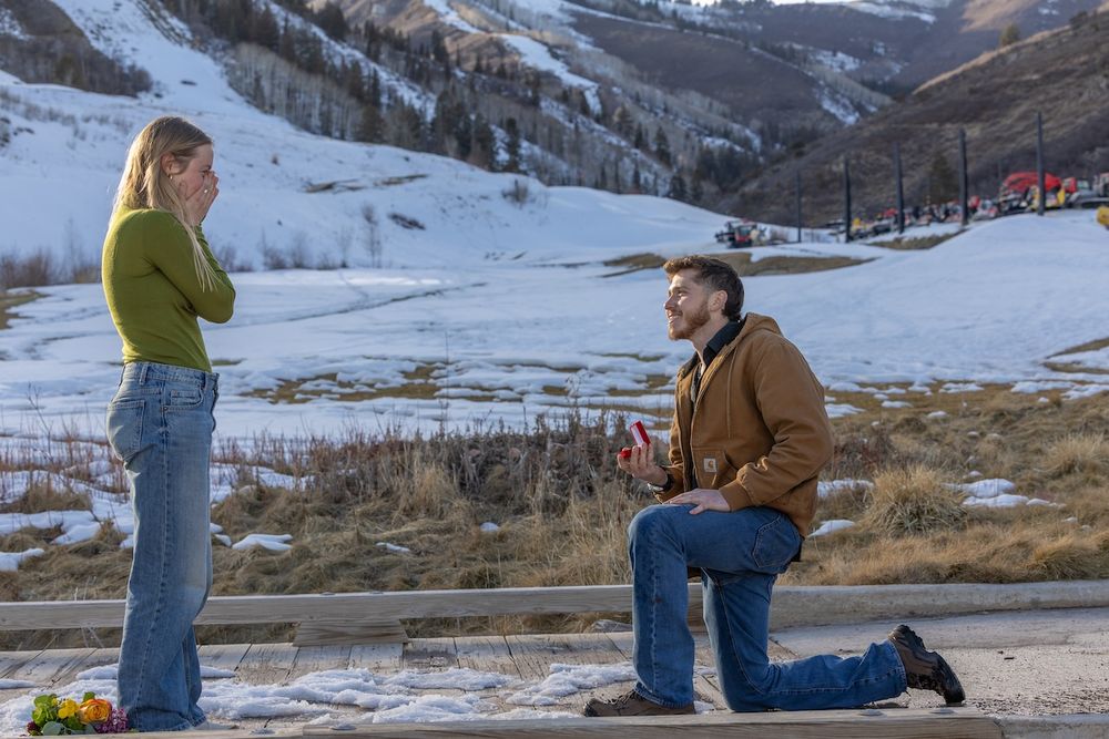 A romantic winter proposal scene unfolds against a snowy mountain backdrop at sunset.