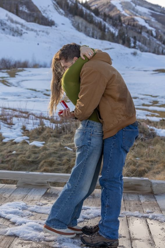 Two people embrace in winter attire against a snowy mountain backdrop on wooden decking.
