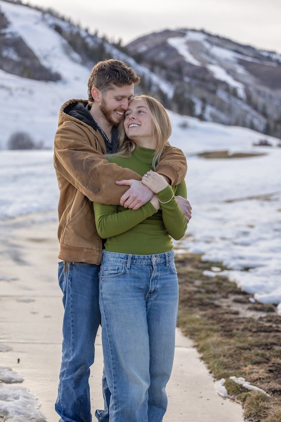 A couple embraces lovingly in a snowy winter mountain landscape wearing casual clothing.