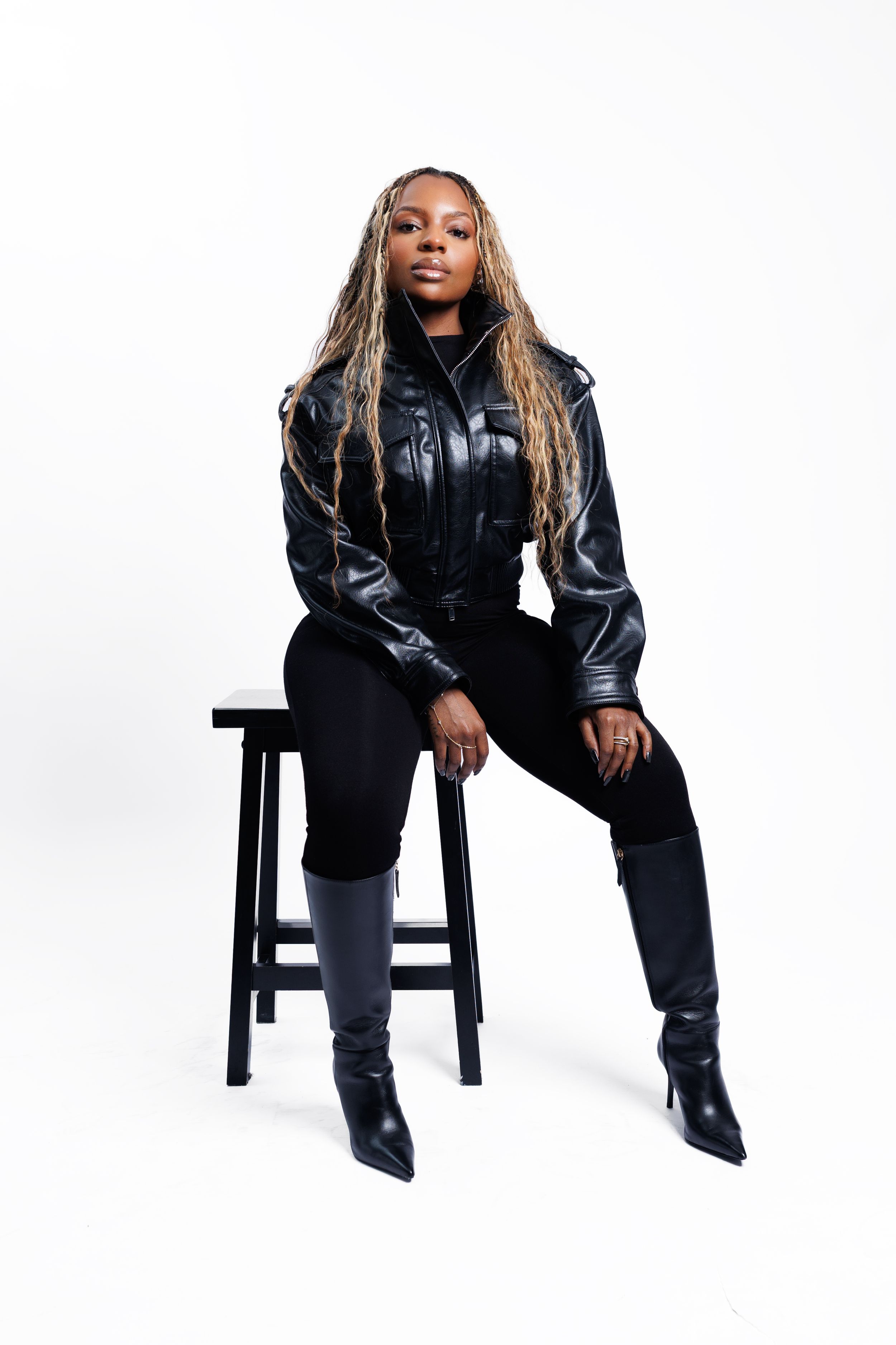 Stylish model in black leather jacket and boots poses confidently on wooden stool against white background.