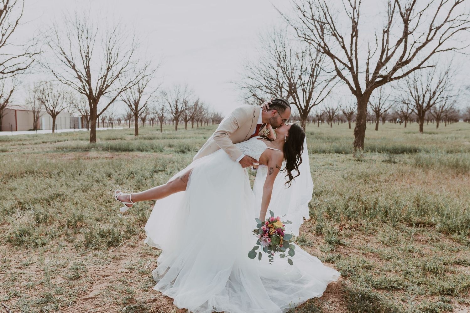 Groom dipping bride in a wedding dress while she holds a colorful bouquet in an orchard.