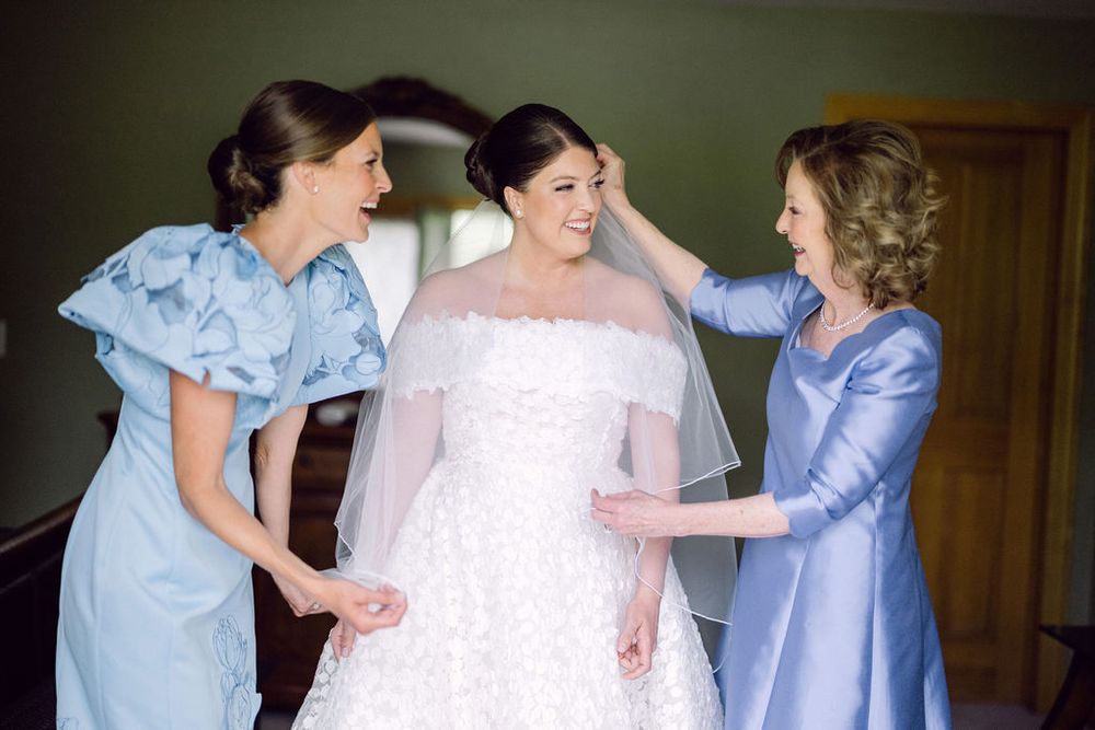Bridesmaids in light blue dresses help adjust the bride's white gown in a sunlit room.