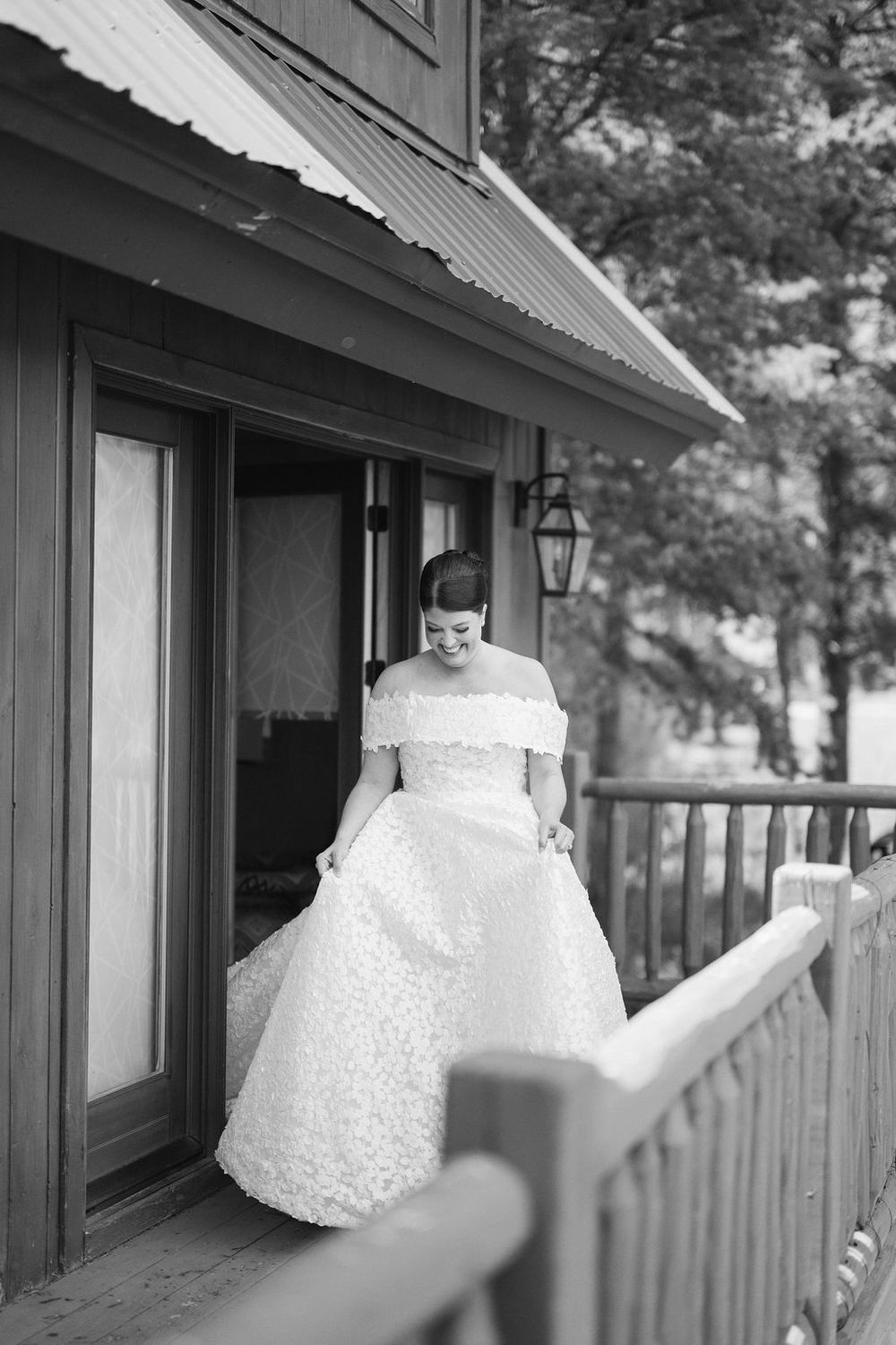 A bride in a white lace off-shoulder wedding gown stands on a rustic wooden porch with a lantern light fixture.
