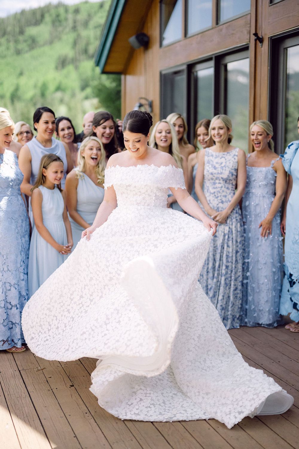 Bride in a white wedding dress twirls on a wooden deck surrounded by bridesmaids wearing light blue floral dresses.