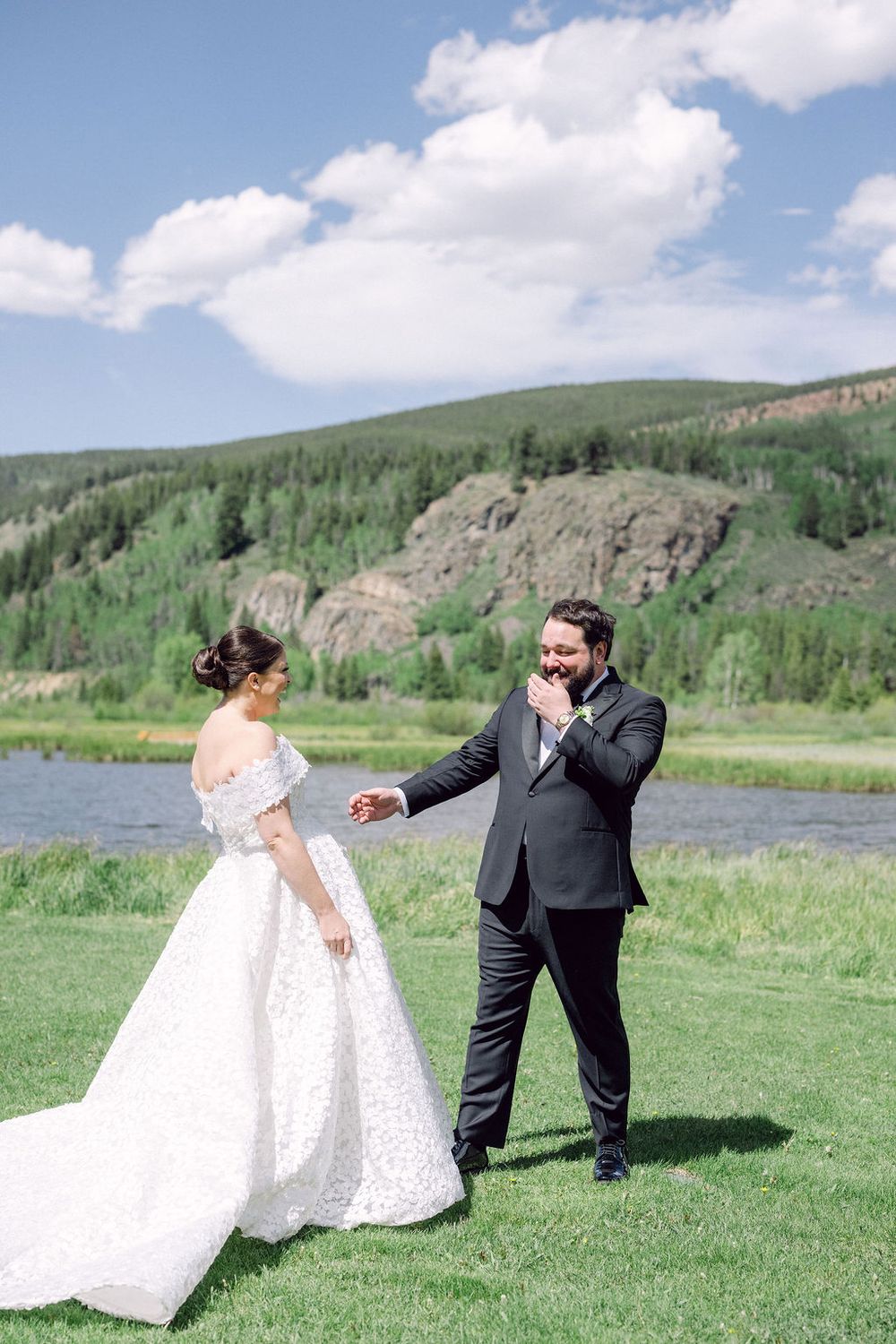 A couple in wedding attire shares playful moments in a scenic mountain meadow by a lake on their special day.