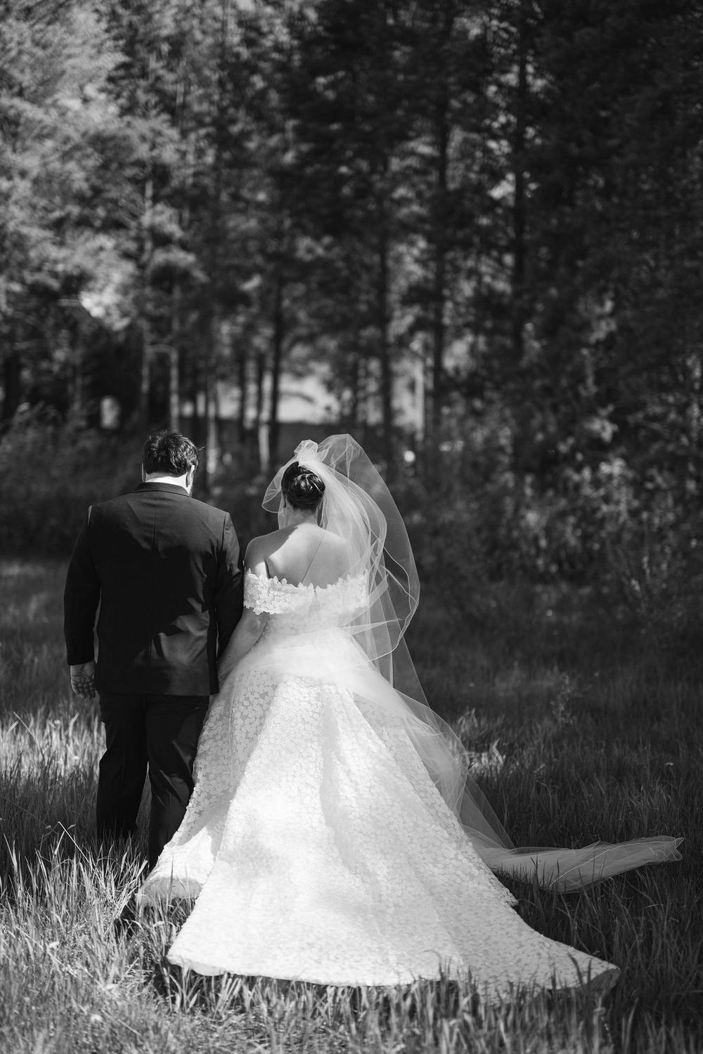 A bride in a flowing white ballgown and veil walks with her partner in a wooded outdoor setting.