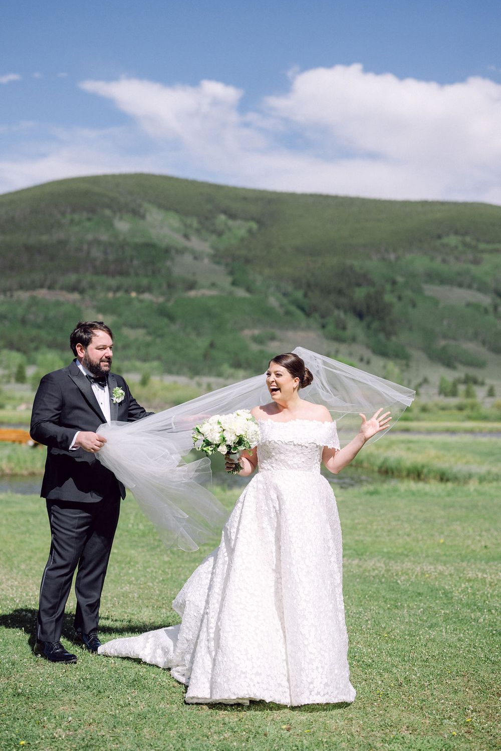 A joyful wedding couple poses in a scenic mountain meadow with the bride's veil flowing in the breeze.