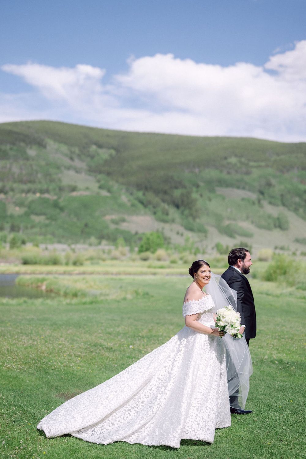 A wedding couple poses in a scenic mountain meadow with rolling green hills and blue skies in the background.