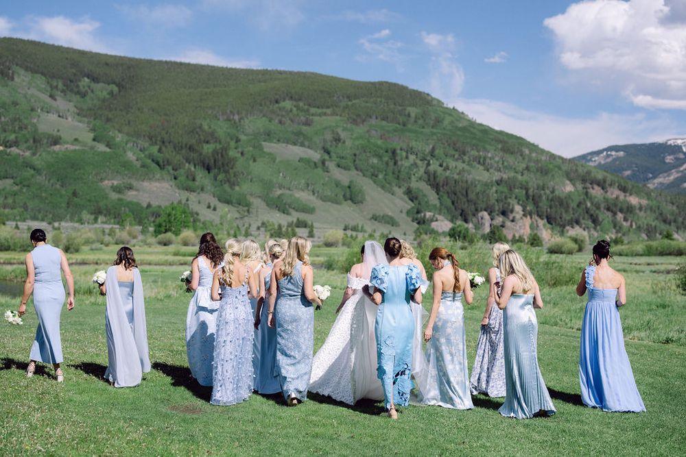 A group in light blue dresses and suits sits in a row on a grassy hillside overlooking a scenic mountain landscape.