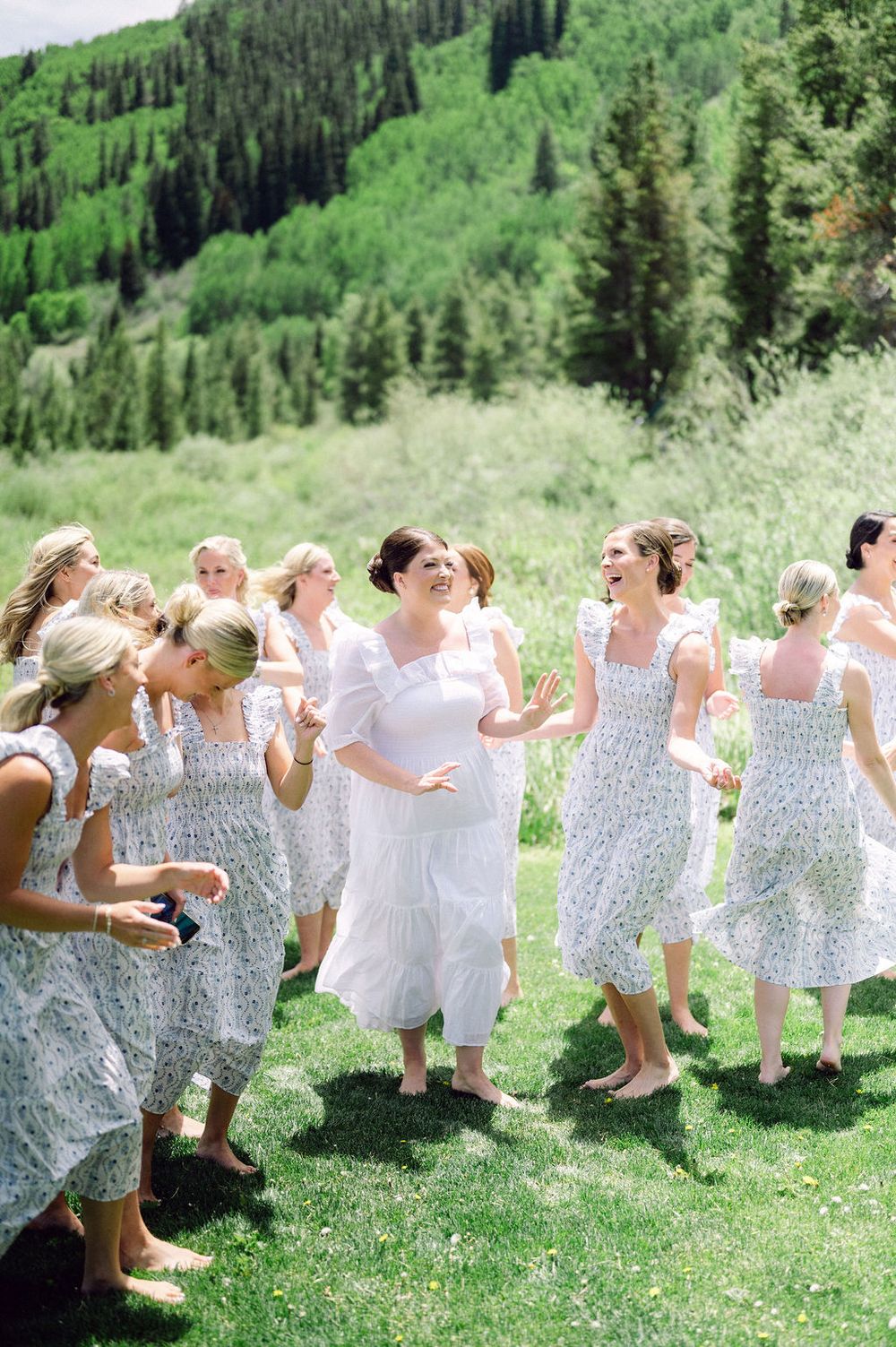 A group in white and floral dresses laugh joyfully together on a grassy mountain meadow surrounded by evergreen forest.