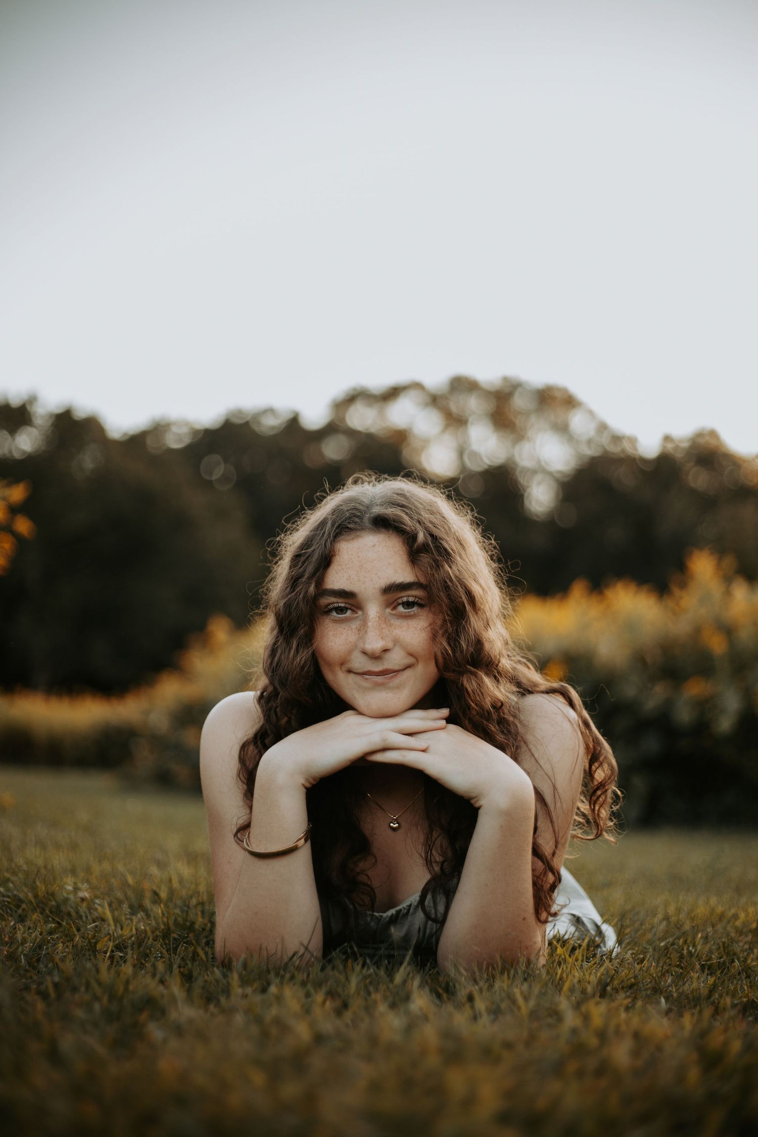 A person with long curly hair lies in a grassy field during golden hour, creating a dreamy outdoor portrait.
