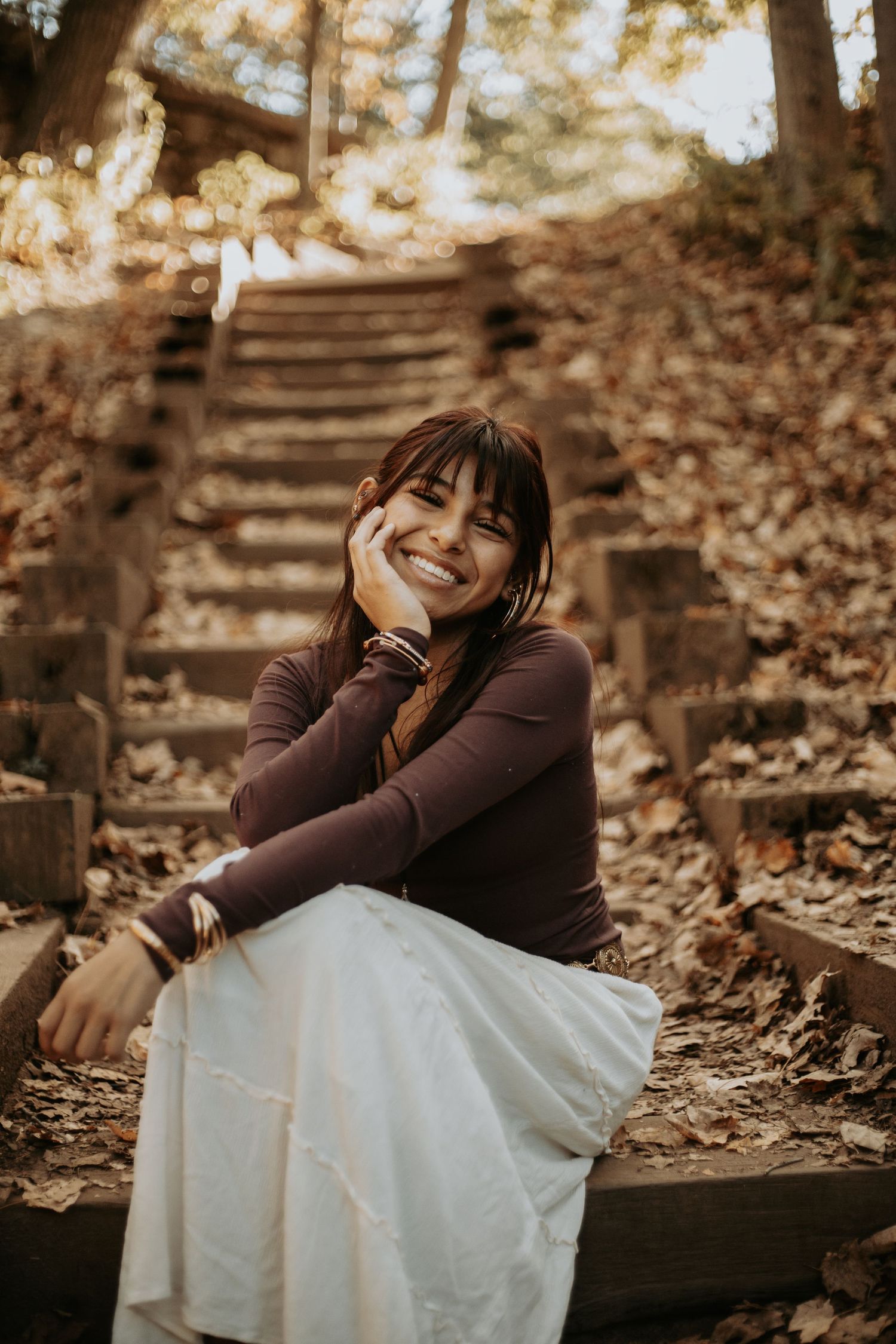 A person in a brown sweater and white dress sits on wooden steps covered in autumn leaves, showing joyful expressions.