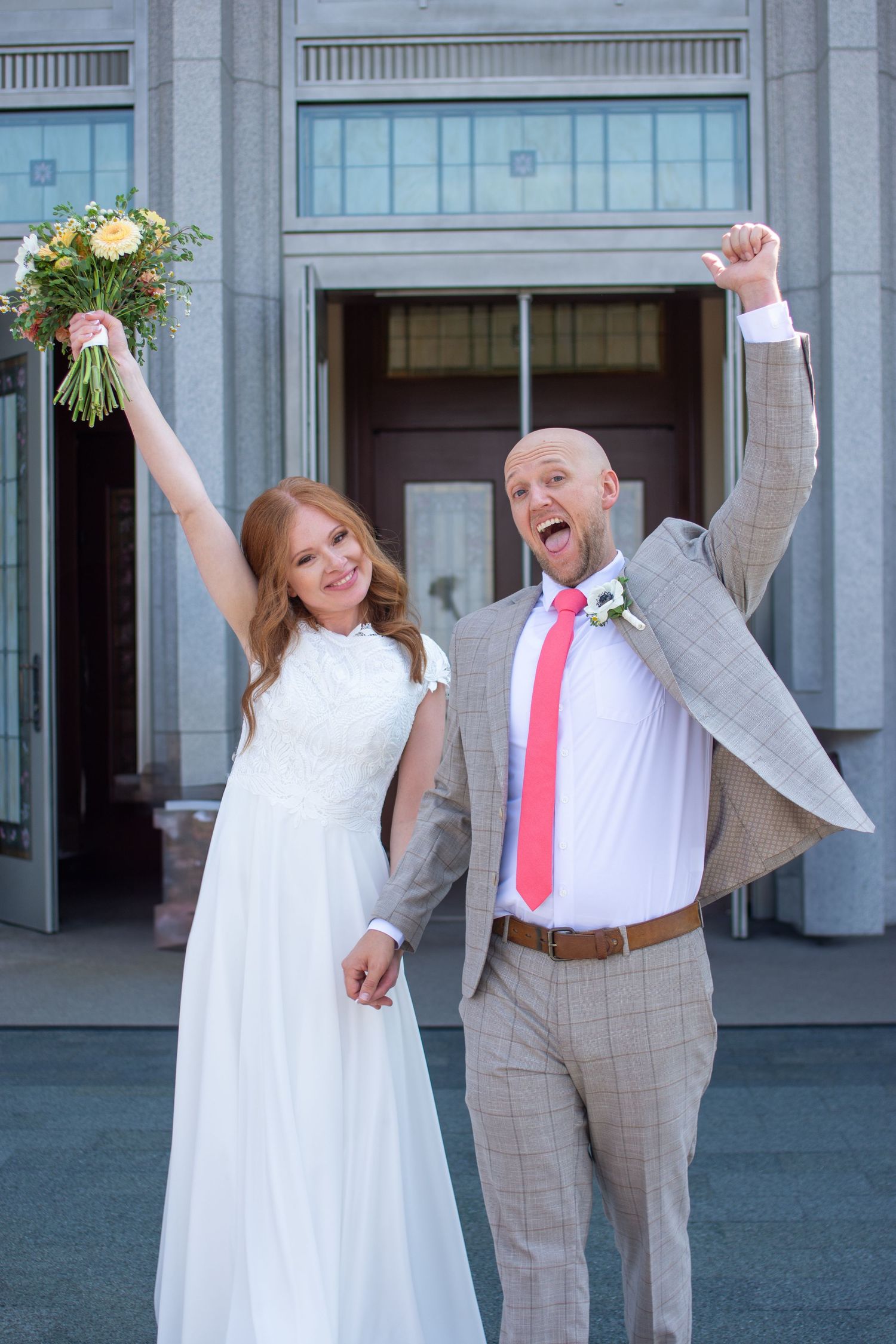 A joyful wedding couple celebrates outside a building, with the bride holding a bouquet and groom wearing a gray suit.