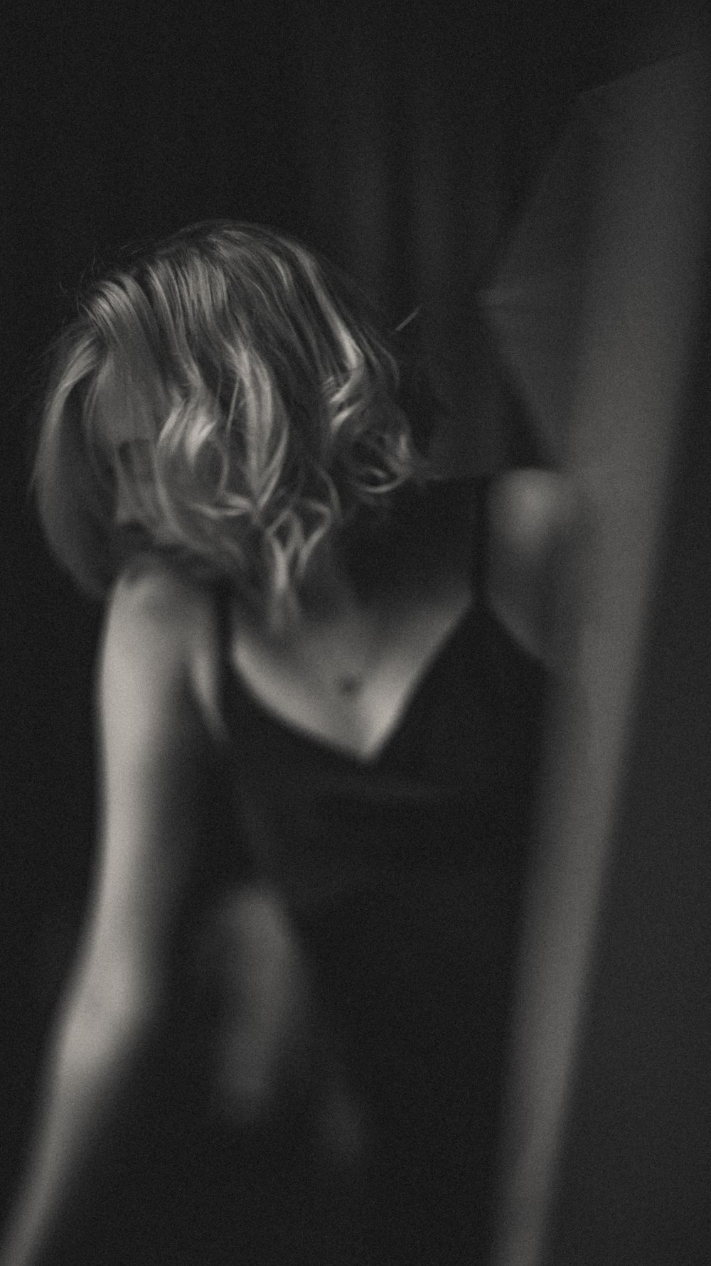 A moody black and white artistic portrait shows a person's hair and shoulder silhouetted against a dark background.