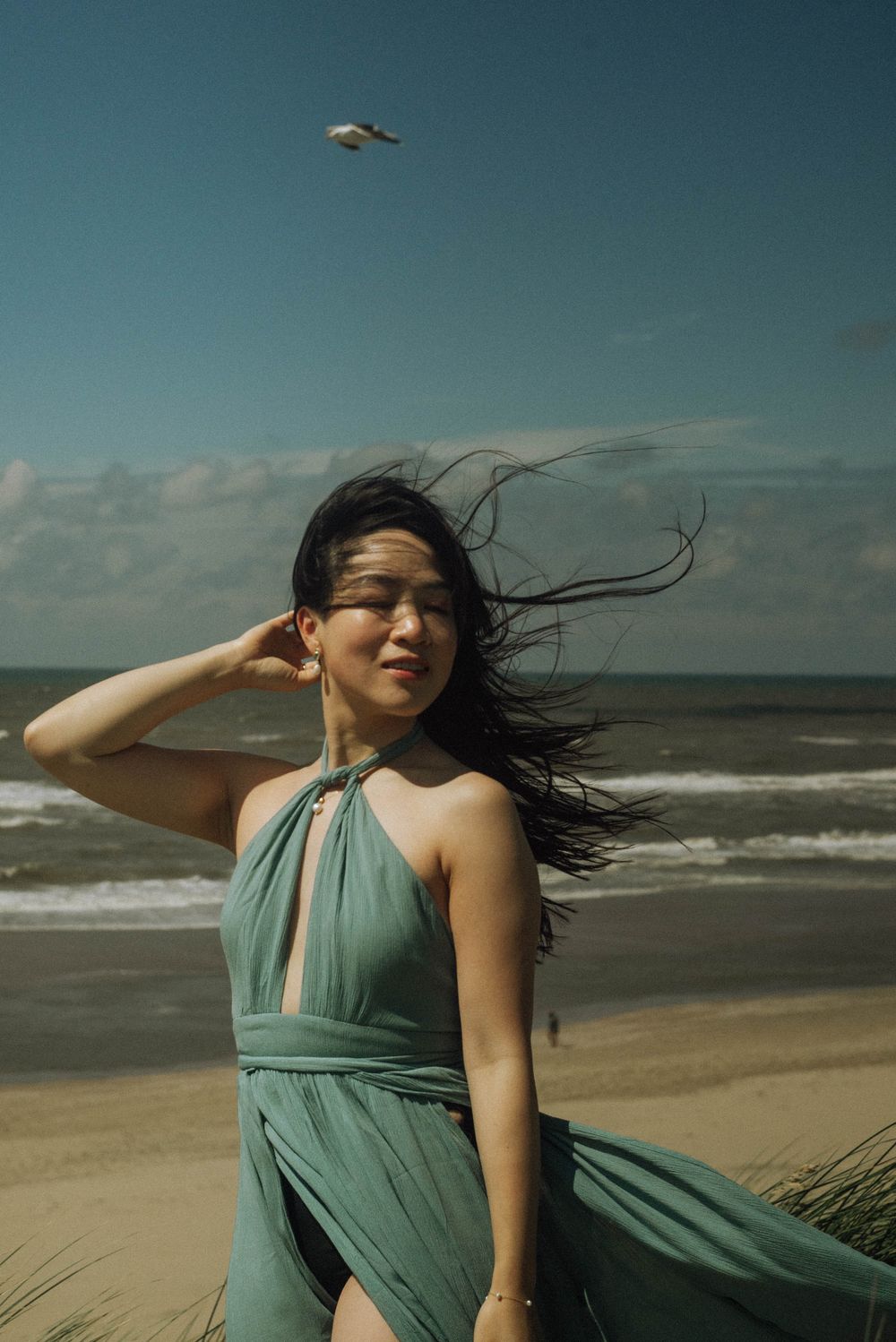A woman in a flowing teal dress stands on a windy beach as her hair and dress billow in the ocean breeze.