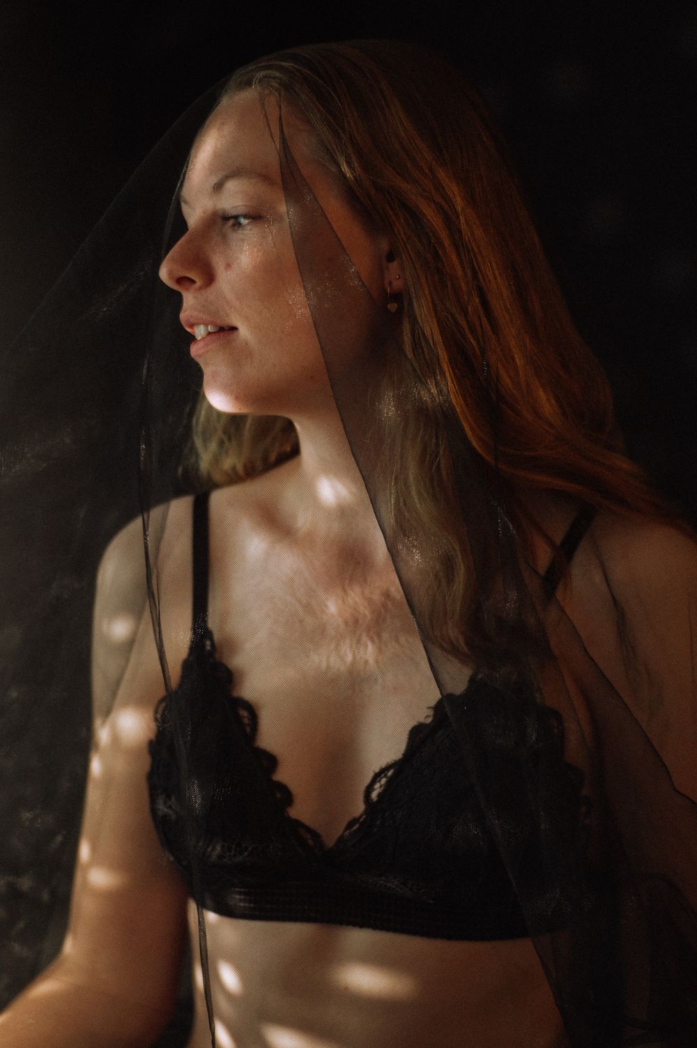 A person with long red hair in a black top poses against a dark background with dramatic lighting and shadows.
