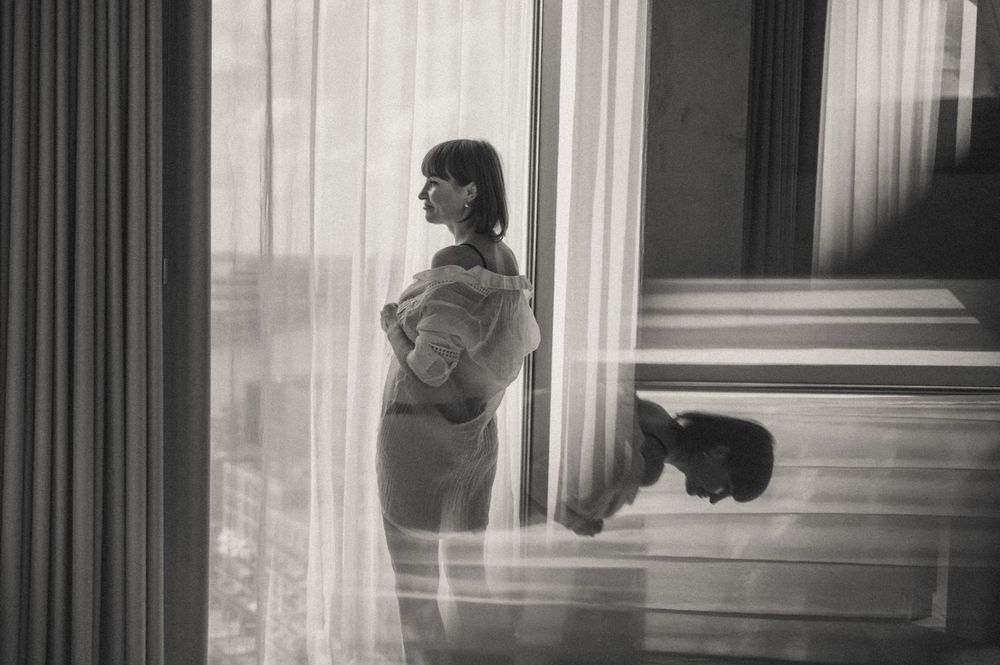 A black and white photo shows a person standing by a window in silhouette, creating a moody atmospheric scene.