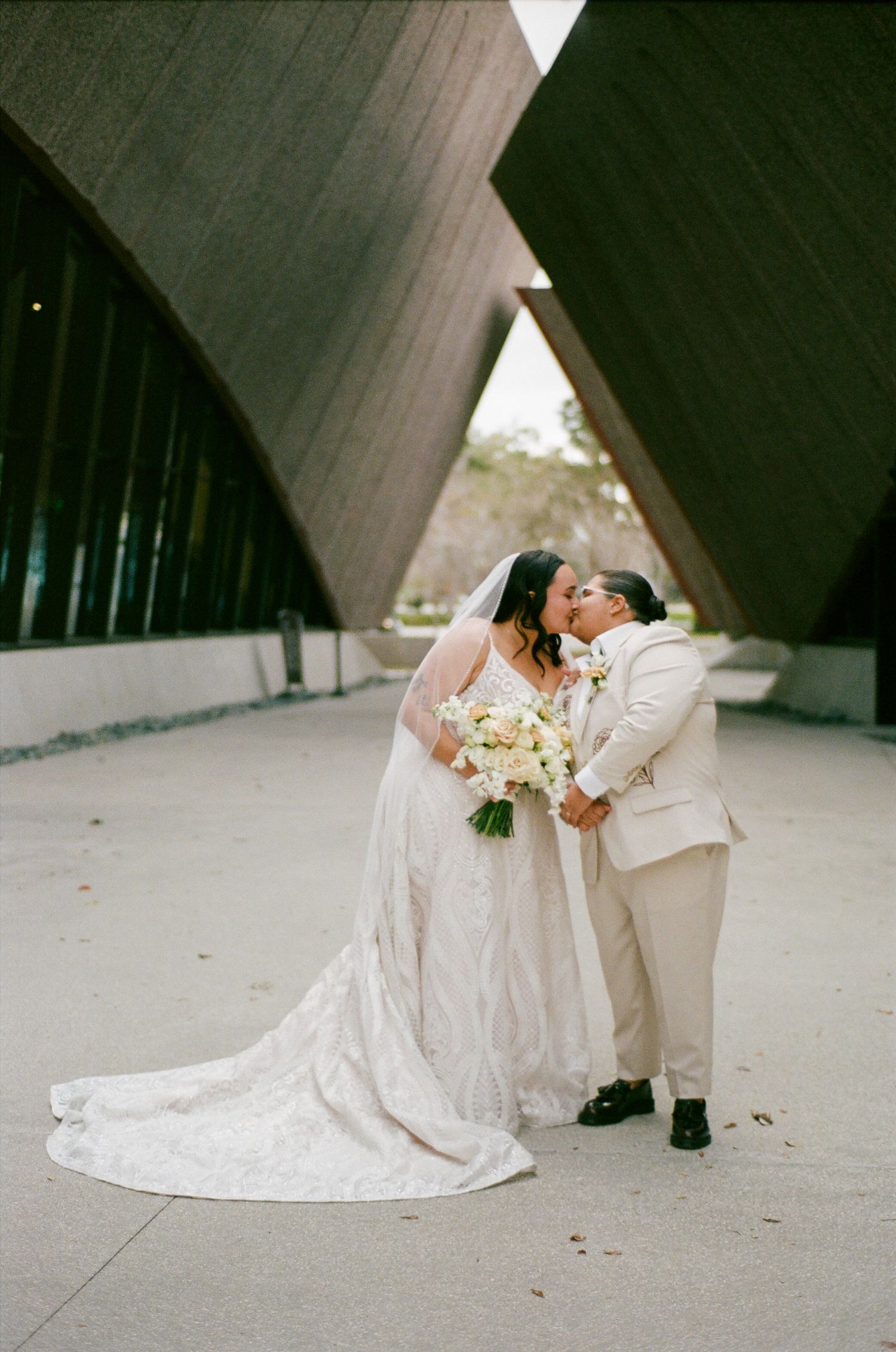 A couple in wedding attire shares an intimate moment on the ground against a concrete architectural backdrop.