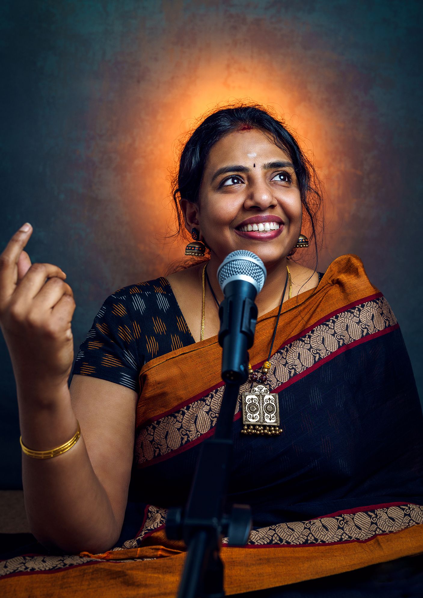 Dramatic lighting captures speaker with microphone in traditional Indian attire against dark background.